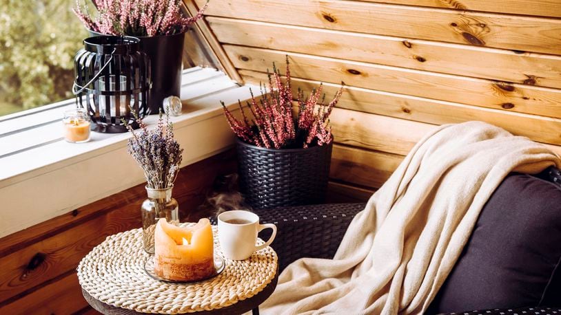 A tiny wooden cabin balcony is seen with heather flowers in a pot, lavender in a bottle vase, a candlelight flame and a soft beige plaid waiting on comfortable garden furniture chair. iSTOCK/COX