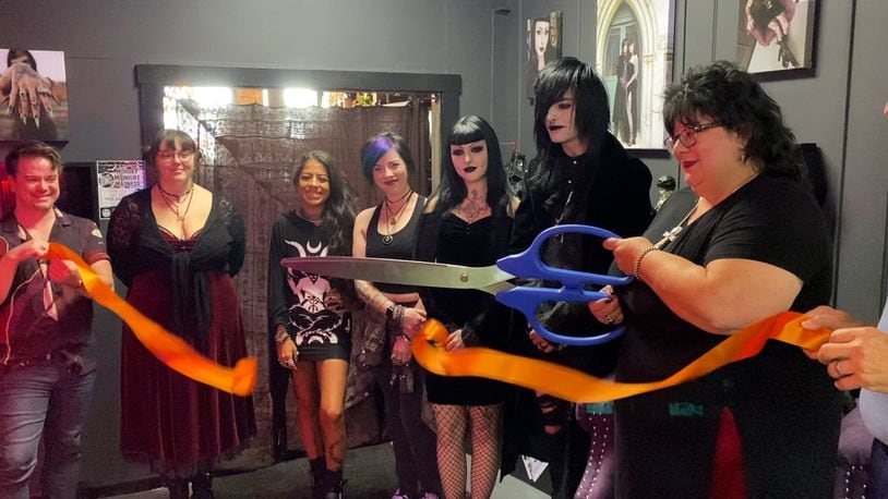 Dark Side Of The Moon is bringing something different to Main Street. The Trad Goth boutique features clothing items and accessories in a goth style. It's located in the back of Seraph By The River, 118 Main St., Hamilton, and both stores are owned by Dawn Singleton. Pictured, with Singleton holding the oversized scissors, is the ribbon cutting of the new boutique on Friday, July 19, 2024. PROVIDED/TVHamilton