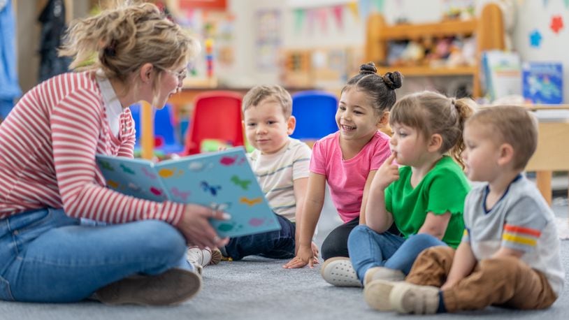 A preschool teacher sits on the floor of her classroom with a small group of students as she reads them a book. FATCAMERA/ISTOCK