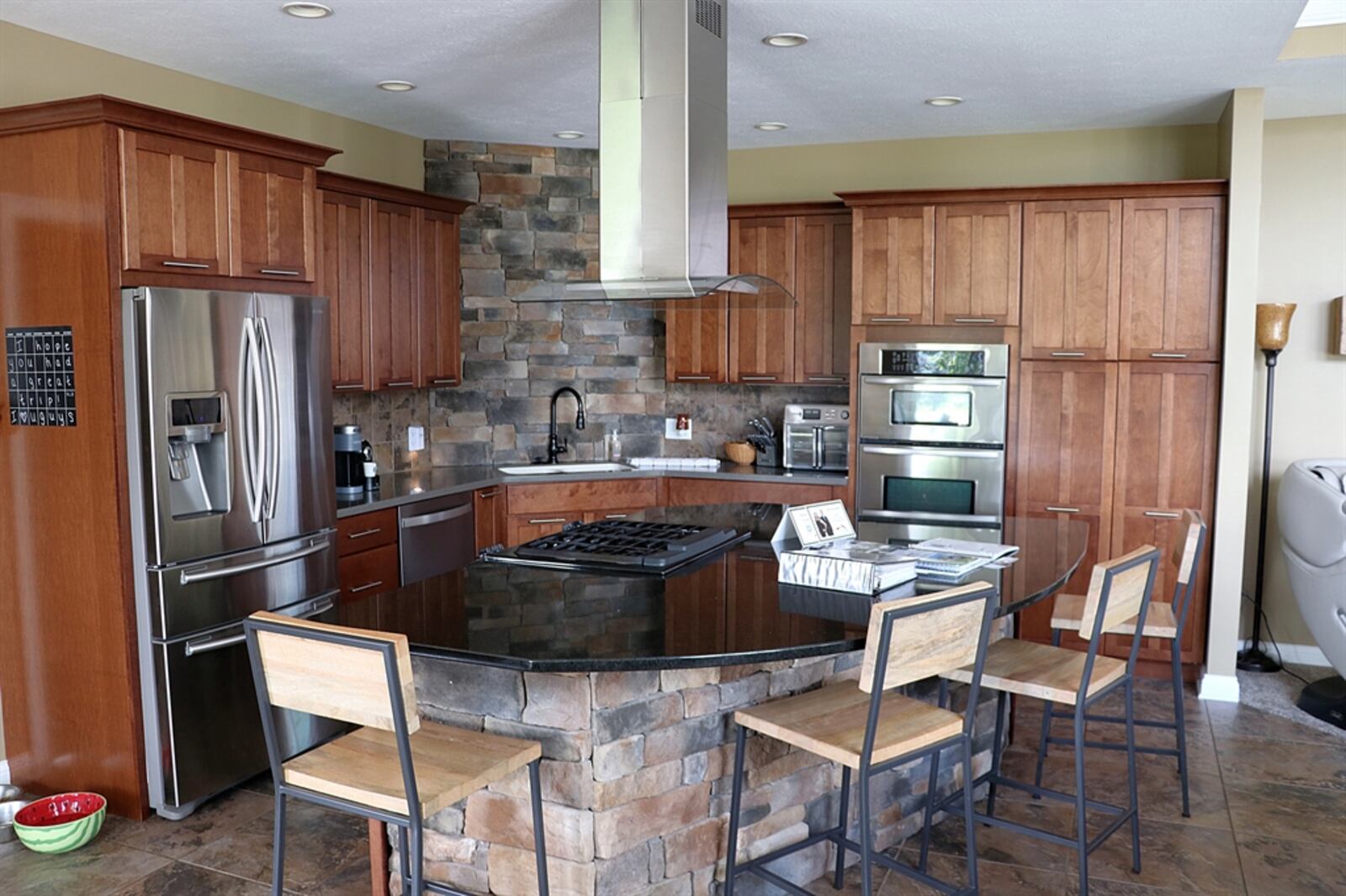 In the kitchen, a large island has stone accents and a gas range within the granite counter. A stainless-steel hood vent hangs above the island.