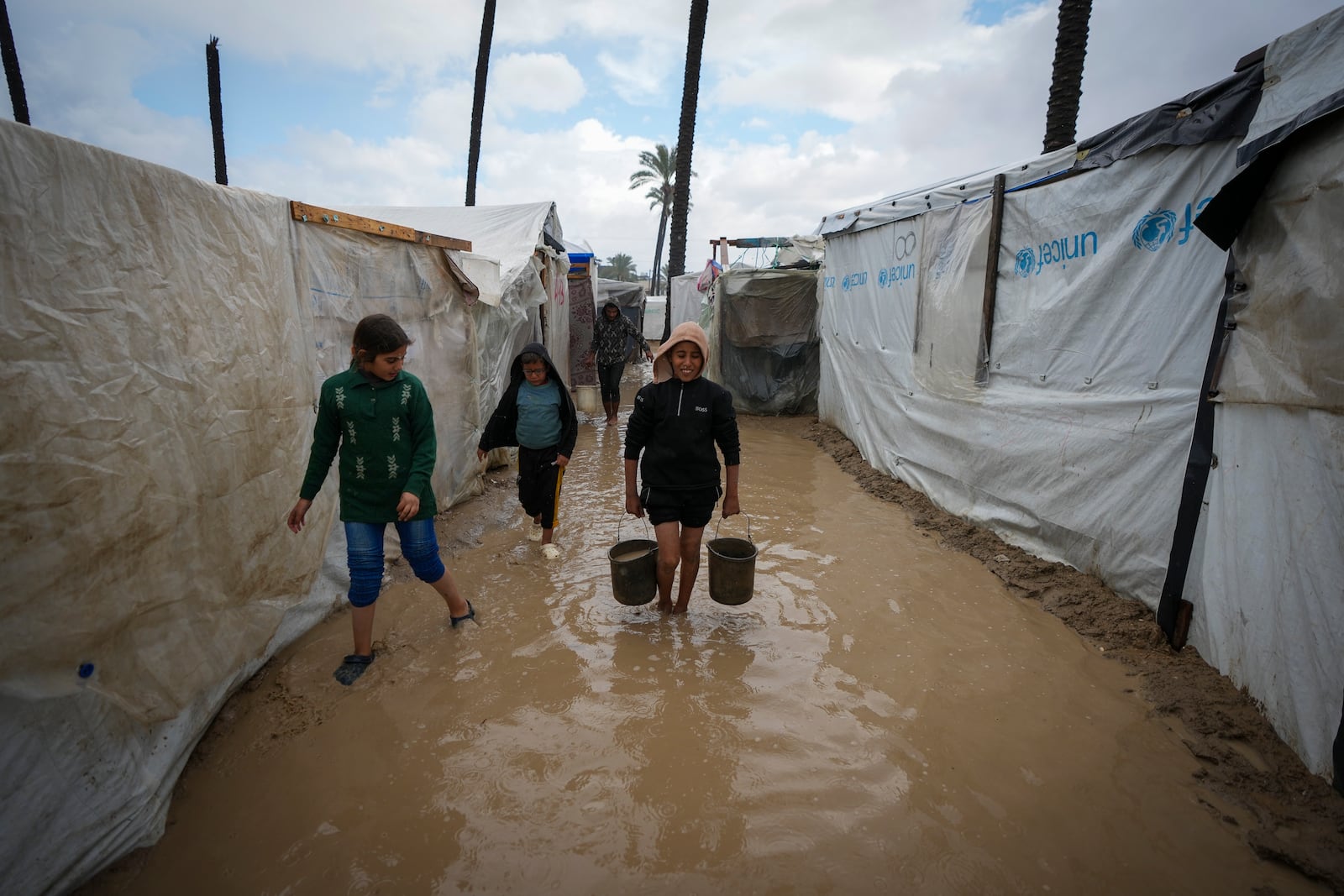 Palestinian children carry buckets of water after overnight rainfall at the refugee tent camp for displaced Palestinians in Deir al-Balah, central Gaza Strip, Tuesday, Dec. 31, 2024. (AP Photo/Abdel Kareem Hana)