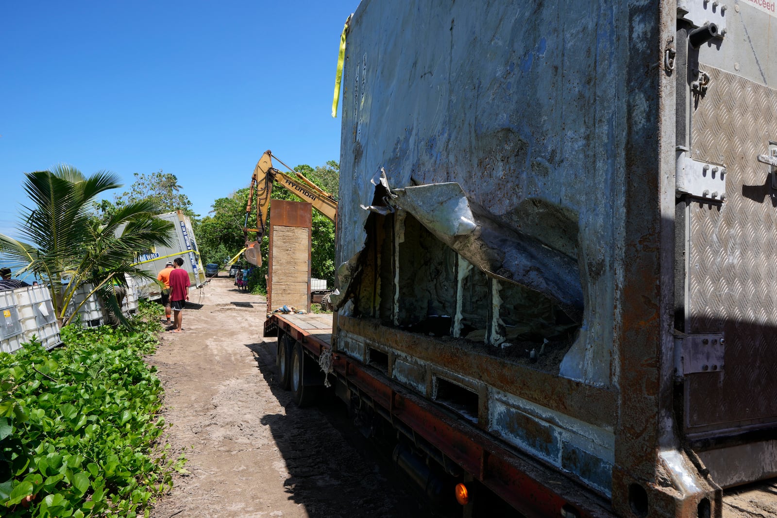 An excavator is used to drag a shipping container onto the beach, left, as another sits on a trailer in the village of Mulivai Safata, Samoa, on Monday, Oct. 21, 2024, near where a New Zealand navy ship ran aground and sank on Oct. 6. (AP Photo/Rick Rycroft)