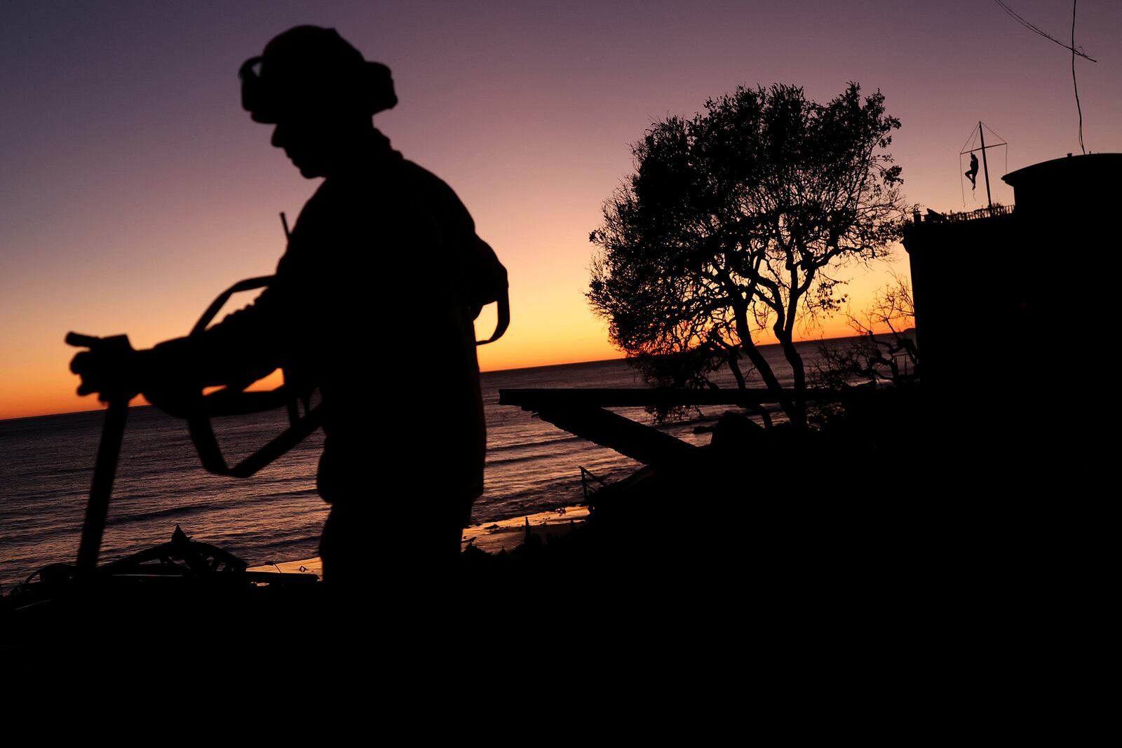 Los Angeles Fire Department's Mike Alvarez works on extinguishing hot spots in the aftermath of the Palisades Fire as a Malibu resident, top right, watches the sunset from atop his beachfront home along the Pacific Coast Highway in Malibu, Calif., Sunday, Jan. 12, 2025. (Scott Strazzante/San Francisco Chronicle via AP)