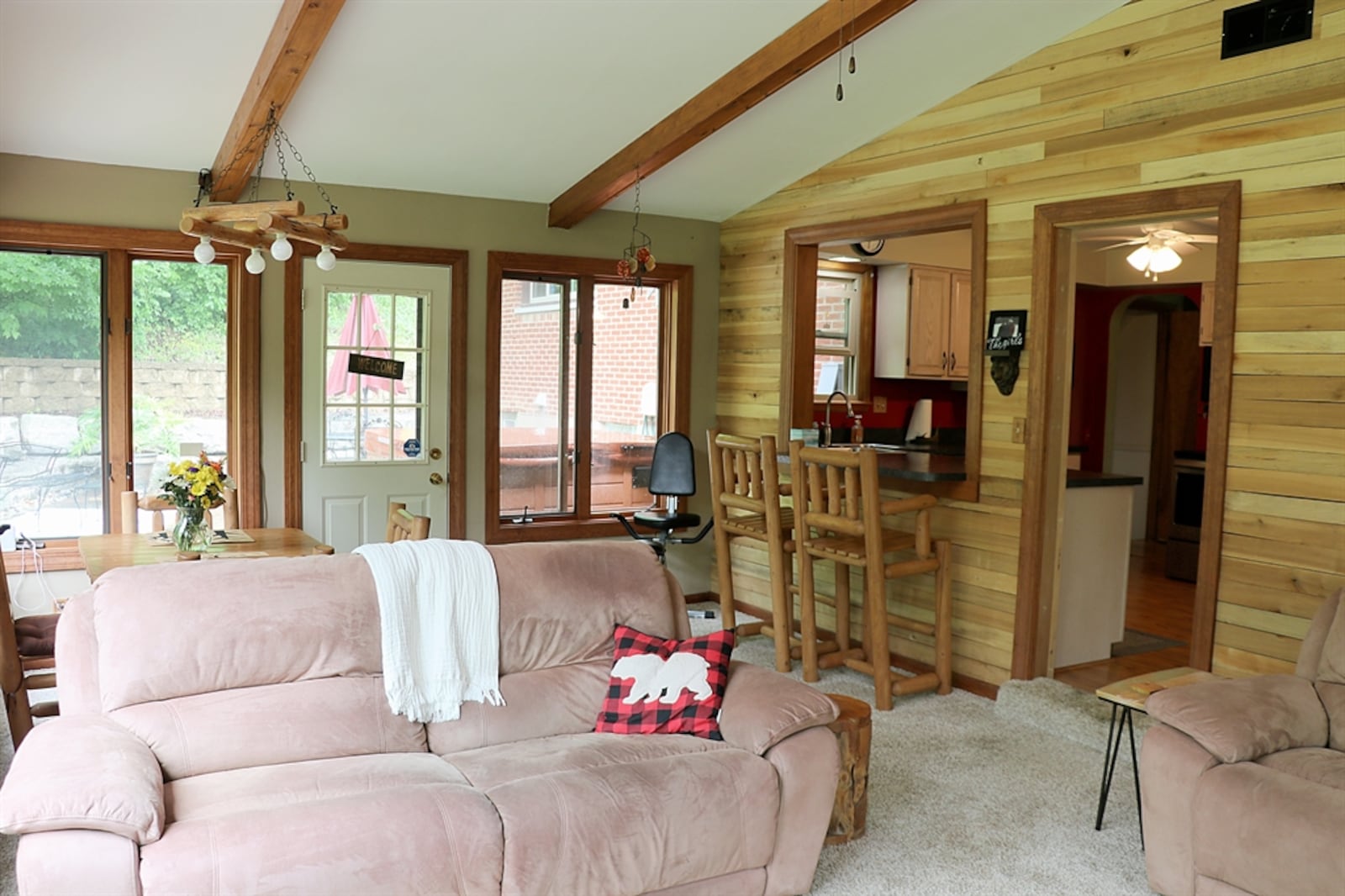 The family room has a cathedral ceiling with triangular windows near the peak. Wood beams accent the ceiling and complement the wood trim around the picture windows. Knotty-pine planks accent another wall and a single door opens out to the courtyard patio. Contributed