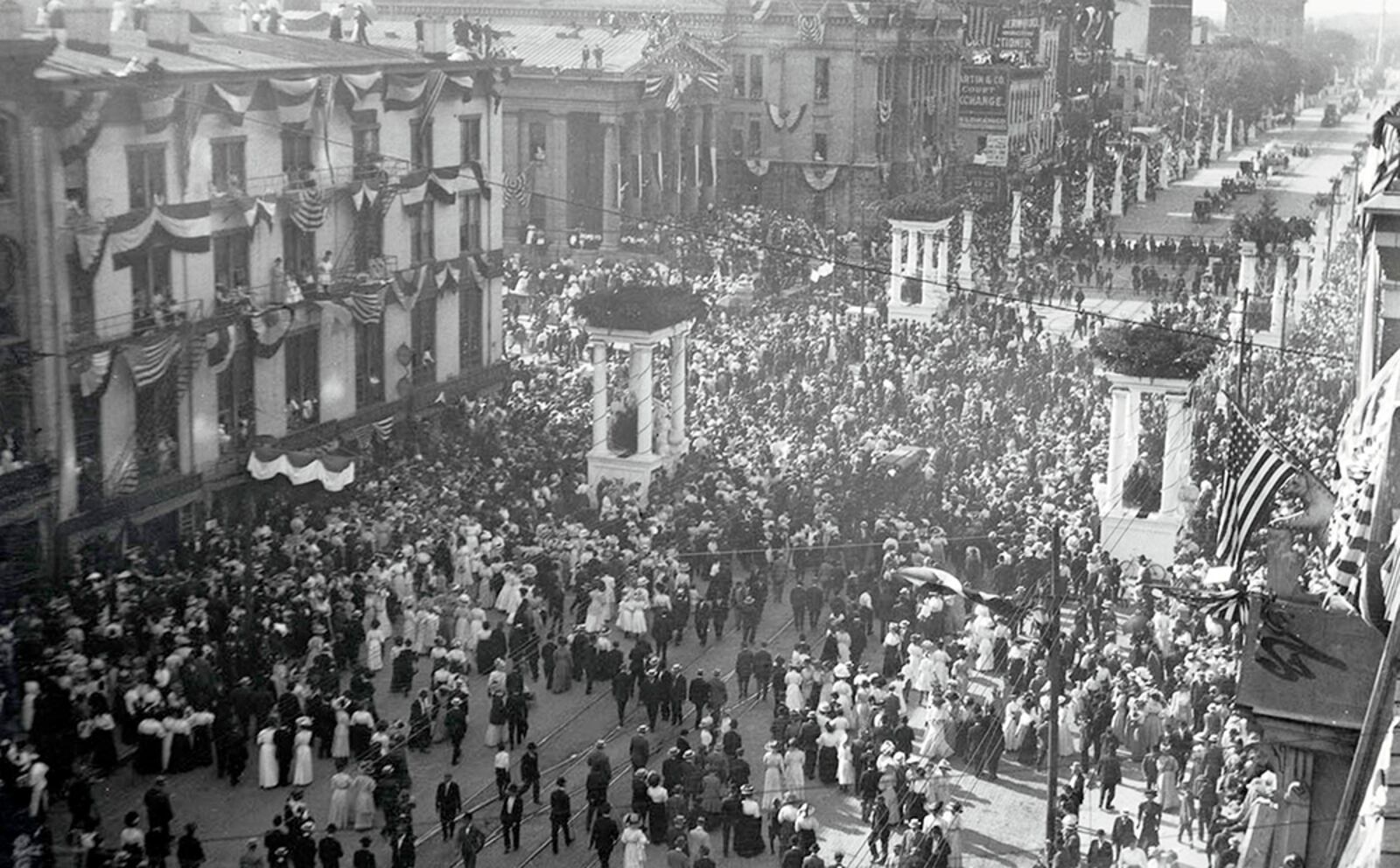 The image shows an overhead view of the Court of Honor located on North Main Street in Dayton, Ohio during the 1909 Wright Brothers Homecoming Celebration. Crowds are walking between the sculptures and columns that line the edge of the street, as the end of the parade approaches the Soldier's Monument. The image shows Main Street looking north from Third Street toward the Soldier's Monument. In the foreground, a polar bear can be seen, as part of the advertisement, above the Ahlers Fur Store located at the Teutonia Bank Building at the northeast corner of Market Street and Main Street. The building on the left side of the image is the Phillips House Co. at the Southwest Corner of Third Street and Main. There is a sign near the corner of this building for Geo. F. Merry Optician at 5 South Main. The Montgomery County Courthouse is located at the top and center of the image and the First Baptist Church is near the upper right corner. Another sign can be seen near the Montgomery County Courthouse for the Chas. E. Underwood Confectioner Company at 29 and 31 North Main Street. The large building, in the upper right corner, is the Harbour Apartments located at 245 North Main Street. Courtesy Special Collections & Archives, Wright State University