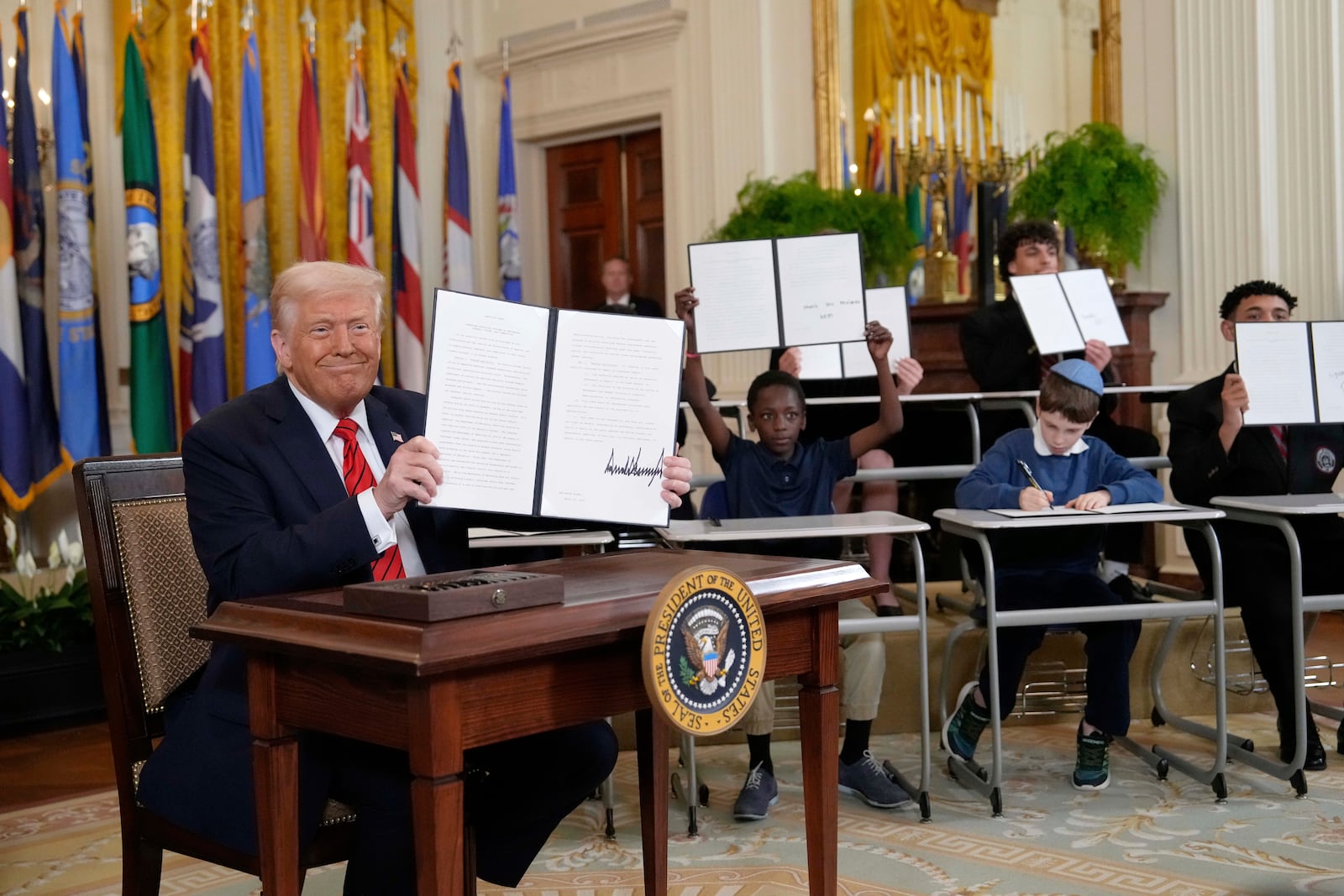 President Donald Trump, left, holds up a signed executive order as young people hold up copies of the executive order they signed at an education event in the East Room of the White House in Washington, Thursday, March 20, 2025. (AP Photo/Ben Curtis)