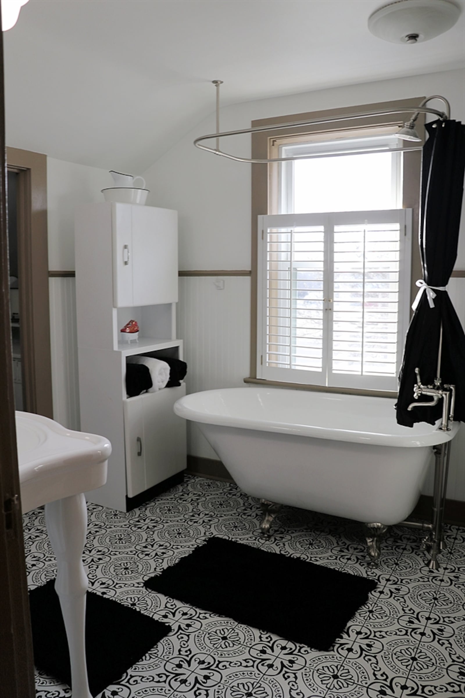 The bath features a claw foot tub with shower right below a window, a porcelain sink with two pedestal legs, ornate ceramic tile flooring and shiplap wood trim. 