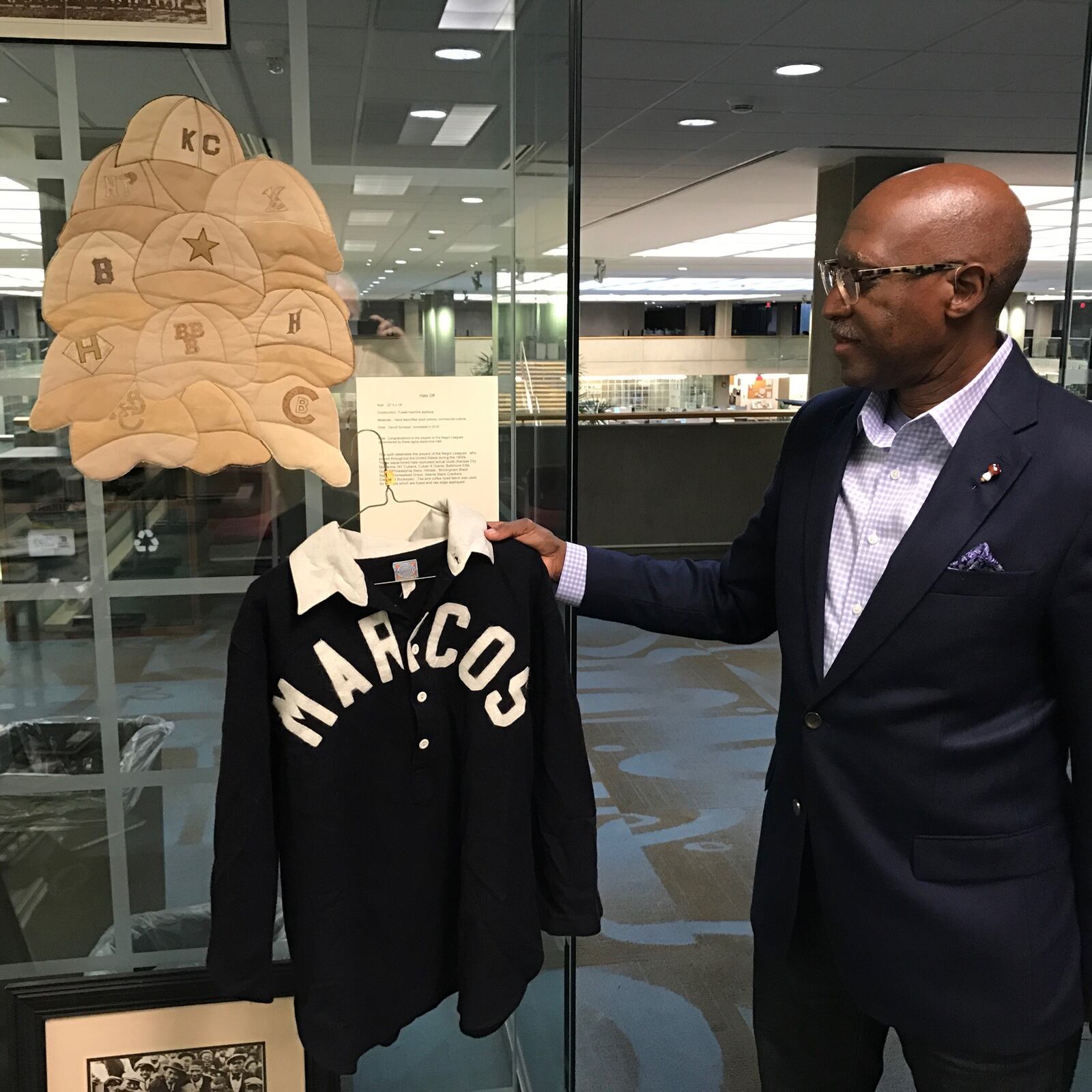 Michael Carter, chief Diversity Officer at Sinclair Community College and former Trotwood Madison and Springfield South High basketball coach,  is the curator of the “Our American Journey exhibit about African American that opens Monday at the school. Included in the exhibit are a pair of quilts made by Kettering’s Carroll Schleppi. depicting Negro Leagues baseball themes. (one is on the wall), Carter is holding a Dayton Marcos jersey. The Marcos were an original Negro Leagues team. Tom Archdeacon/STAFF