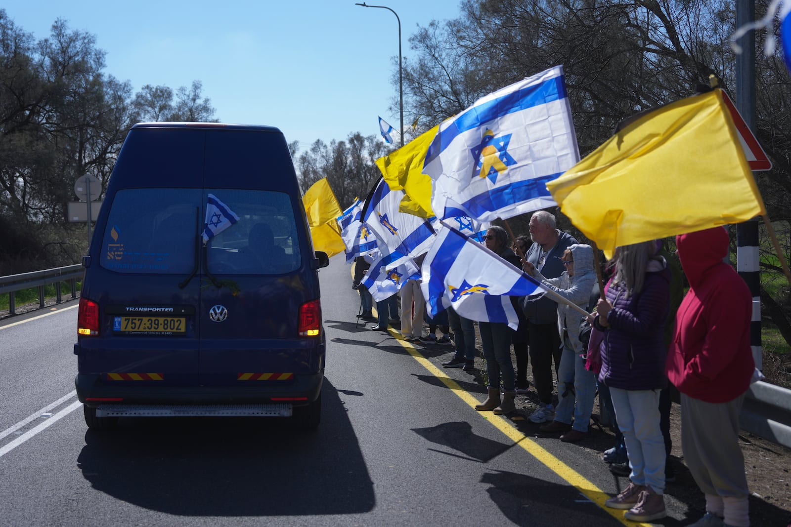 Mourners wave Israeli flags as the car carrying the coffin of former Israeli hostage Oded Lifshitz passes by at the entrance of Kibbutz Nir Oz, where his funeral will take place, in southern Israel, on Tuesday, Feb. 25, 2025. Lifshitz was abducted by Hamas on Oct. 7, 2023, and his remains were returned from Gaza to Israel last week as part of a ceasefire with Hamas. (AP Photo/Ohad Zwigenberg)