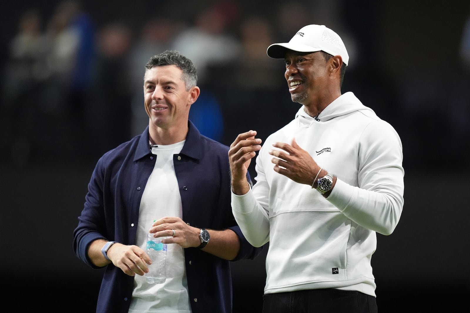 Rory McIlroy and Tiger Woods stand on the green as they watch New York Golf Club and The Bay Golf Club warmup for the inaugural match of the TMRW Golf League, Tuesday, Jan. 7, 2025, in Palm Beach Gardens, Fla. TGL features six teams of four players competing against each other in a tech-infused arena the size of a football field. (AP Photo/Rebecca Blackwell)