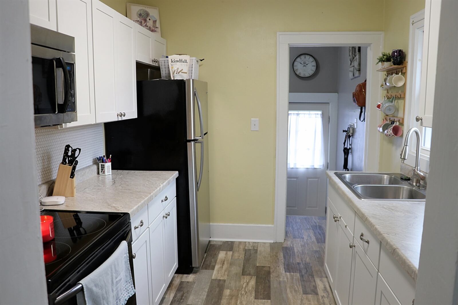 White cabinetry, with light-gray, solid-surface counters, complements the gray wood-vinyl flooring of the kitchen. Two walls are filled with the cabinetry and counters. There is a double-sink below a window, and the kitchen comes equipped with a microwave.