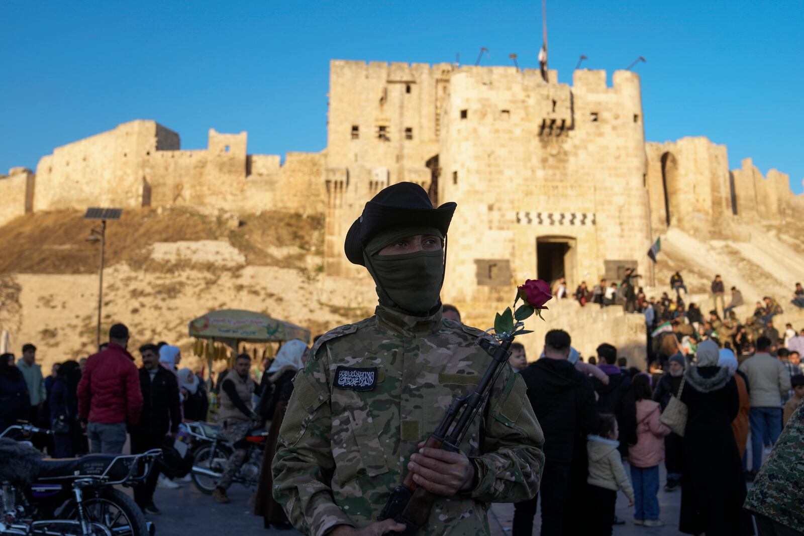 A Syrian fighter guards holding a gun with a flower placed in the barrel, as residents visit the ancient Aleppo Citadel in the old city of Aleppo, Syria, Saturday, Dec. 14, 2024. (AP Photo/Khalil Hamra)