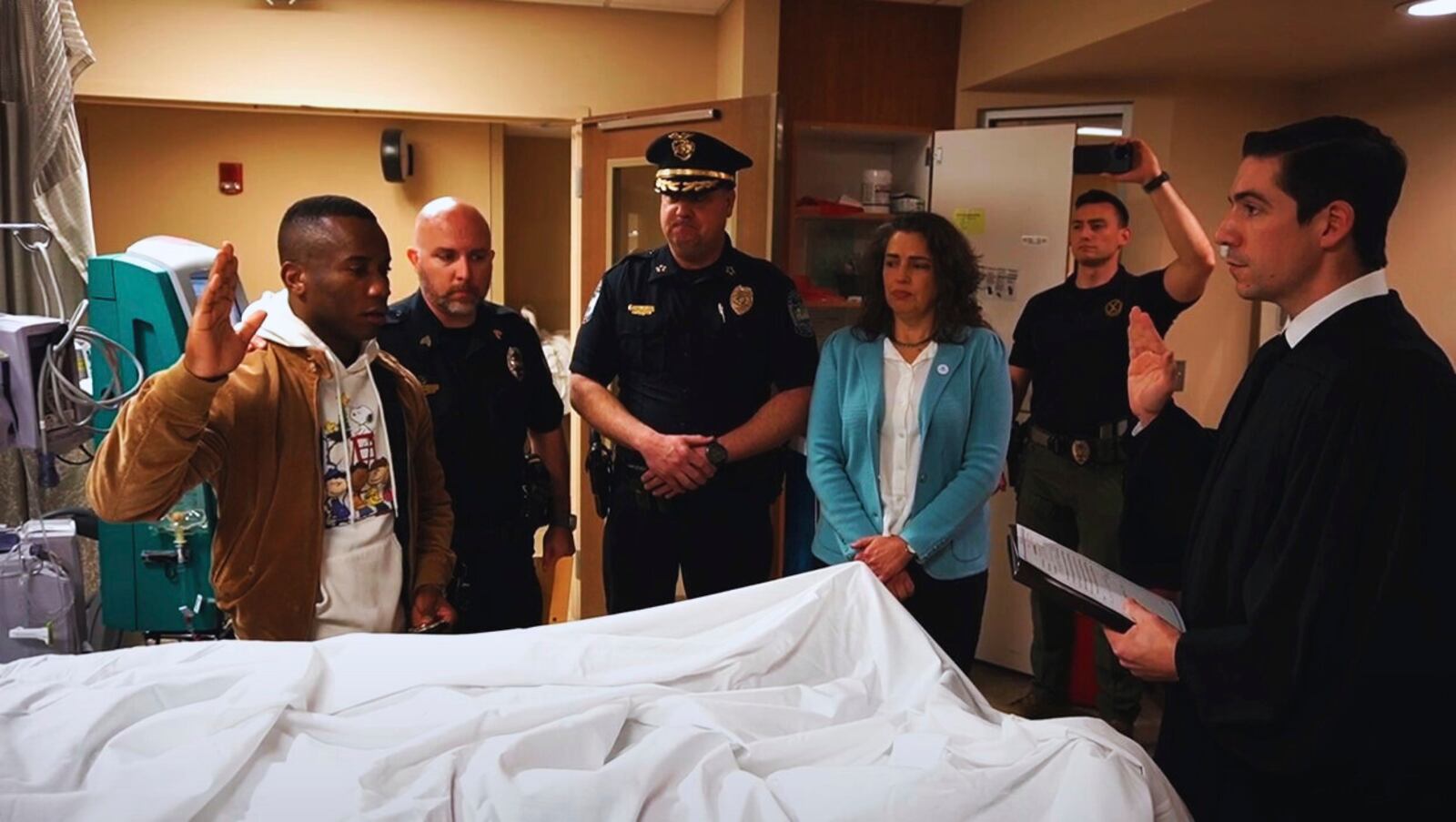 This undated photo provided by the Knoxville, Tenn., Police Department shows Tristants Simpson, a fellow recruit class member, taking the police oath on behalf of Wisbens Antoine, while Sgt. Jimmy Wilson, Police Chief Paul Noel, Mayor Indya Kincannon, Officer Terry Crowe and Municipal Court Judge Tyler Caviness, look on. (Scott Erland/Knoxville Police Department via AP)