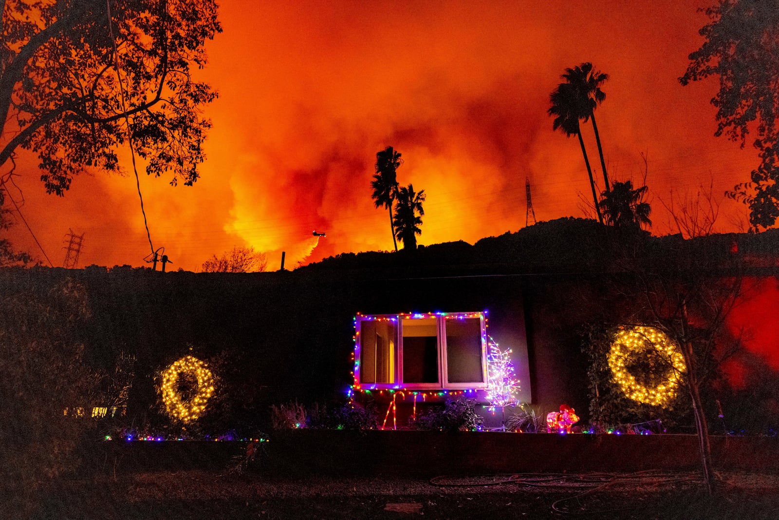 A helicopter drops water on the Palisades Fire behind a home with Christmas lights in Mandeville Canyon, Friday, Jan. 10, 2025, in Los Angeles. (AP Photo/Ethan Swope)