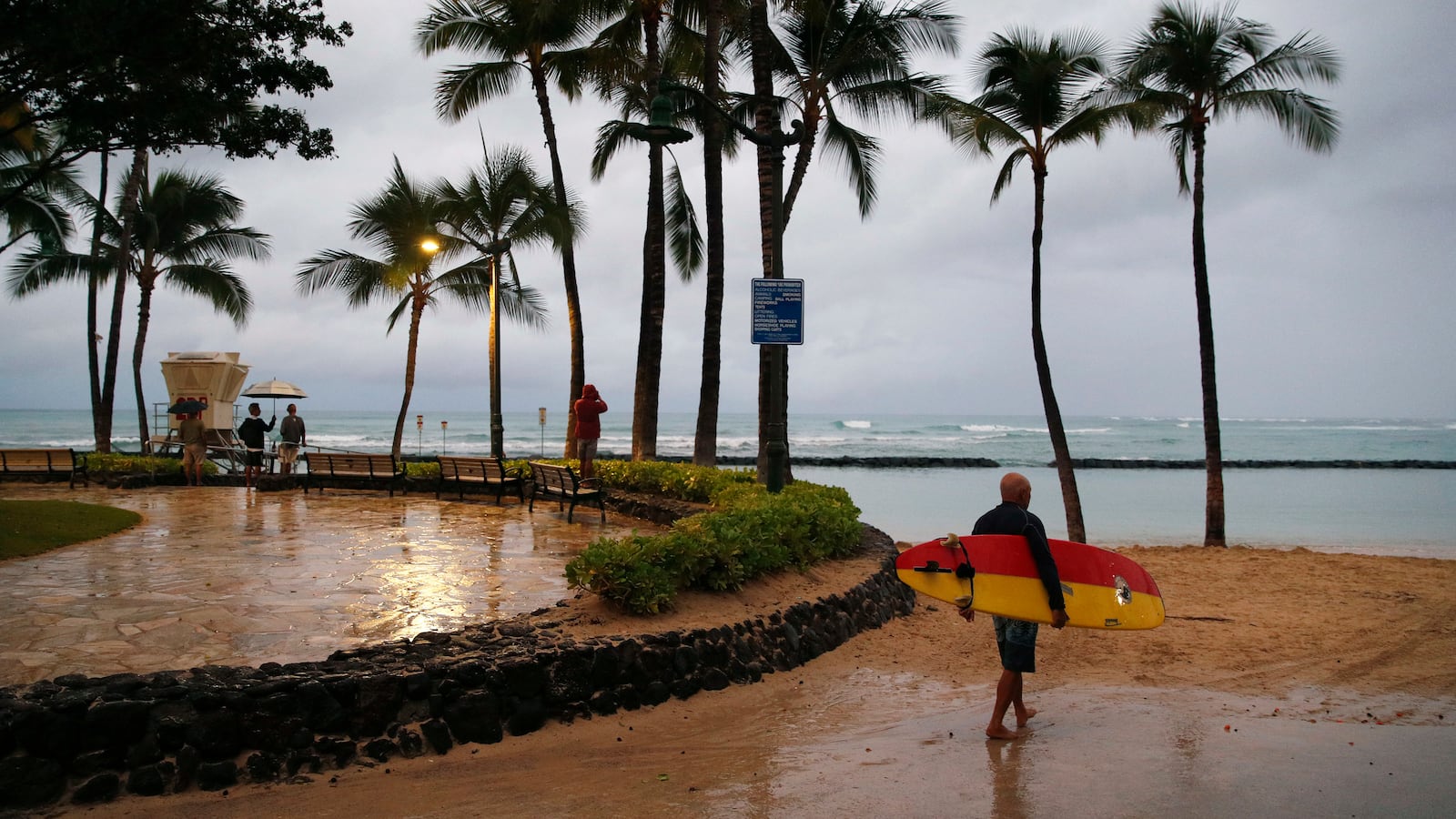 A surfer walks along Waikiki Beach in a light rain from Tropical Storm Lane, Saturday, Aug. 25, 2018, in Honolulu.