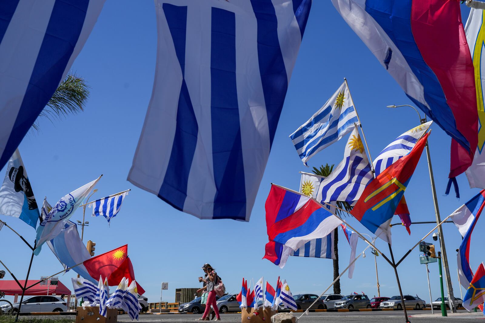 Pedestrians pass by Uruguay's national flag and political party banners for sale on the day of the presidential run-off election in Montevideo, Uruguay, Sunday, Nov. 24, 2024. (AP Photo/Natacha Pisarenko)