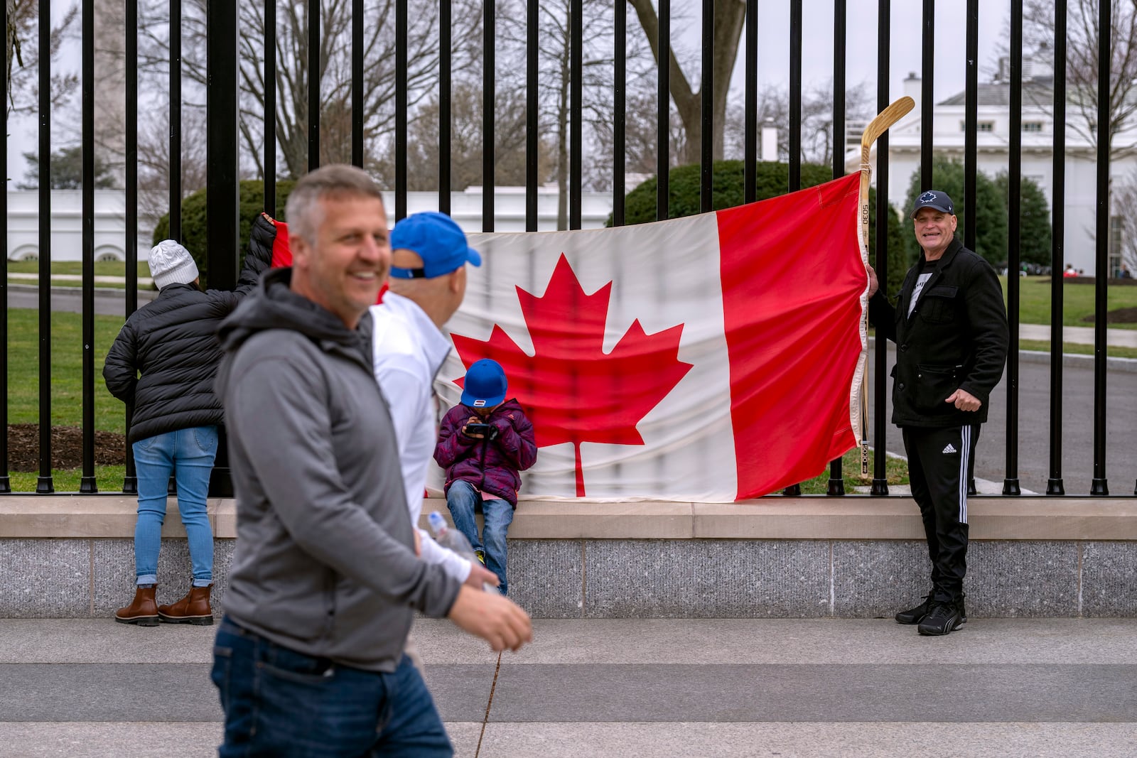 Pedestrians walk past as Toronto residents Douglas Bloomfield, from right, his son Phoenix and wife Ame, left, who are on vacation in Washington from Toronto, hold a Canadian flag and an ice hockey stick to show their support for Canada regarding trade tariffs, in front of the White House in Washington, Thursday, March 13, 2025. (AP Photo/Ben Curtis)