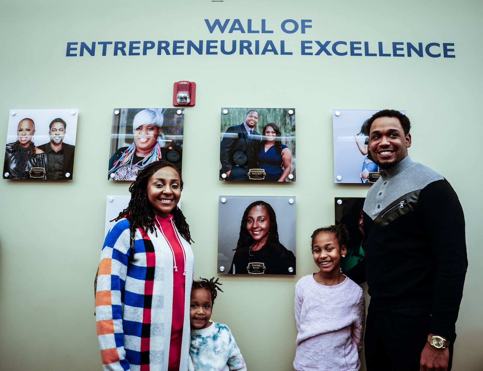 Courtney and Joshua Barrett and their daughters, Bailey, 4, and Brooklyn, 9, get their pictures taken in front of the Entrepreneurial Wall of Excellence where Courtney's photograph is displayed. Courtney founded Just Cakin' It LLC. JIM NOELKER/STAFF