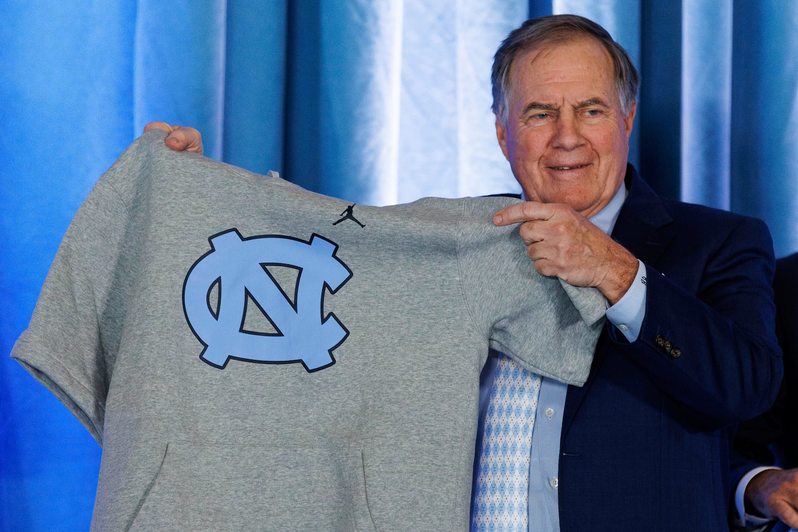 FILE - New North Carolina football coach Bill Belichick holds up a UNC branded sleeveless hoodie presented to him during an NCAA college football news conference announcing his hiring, Dec. 12, 2024, in Chapel Hill, N.C. (AP Photo/Ben McKeown, File)