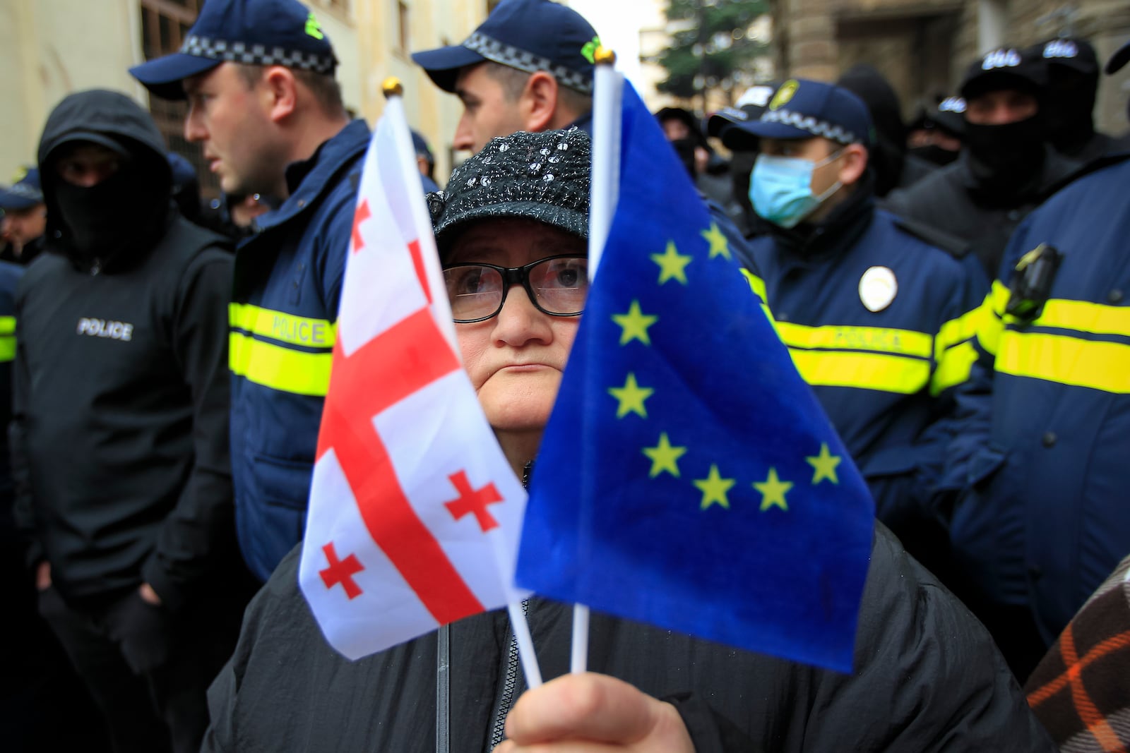 A woman with a EU and a Georgian national flags stands in front of police blocking a street during a rally to demand new parliamentary elections in the country, near the Parliament's building in Tbilisi, Georgia, on Monday, Nov. 25, 2024. (AP Photo/Shakh Aivazov)