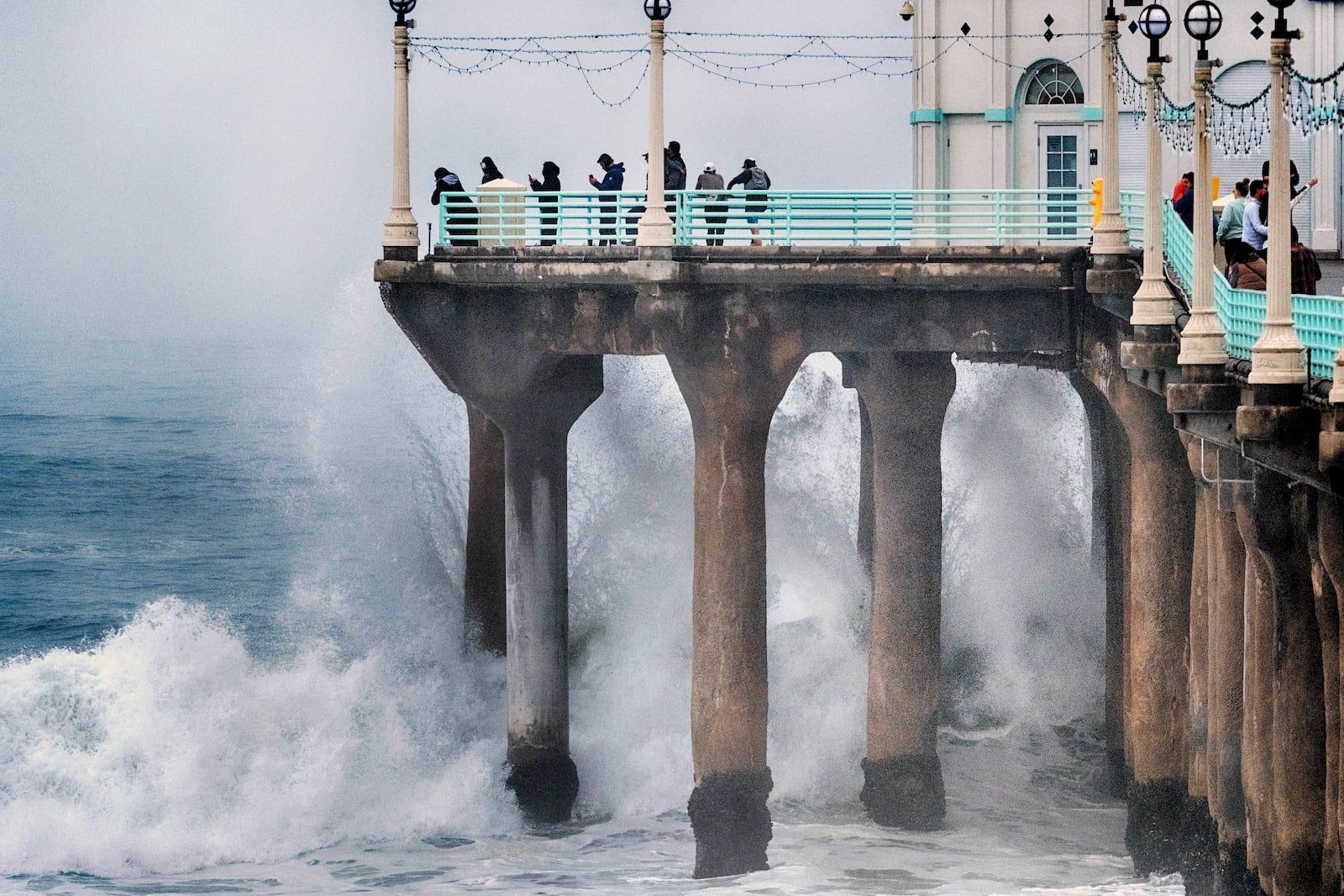 People stand at the end of the Manhattan Beach Pier and watch high surf pound the pylons on Tuesday, Dec. 24, 2024 in Manhattan Beach, Calif. (AP Photo/Richard Vogel)