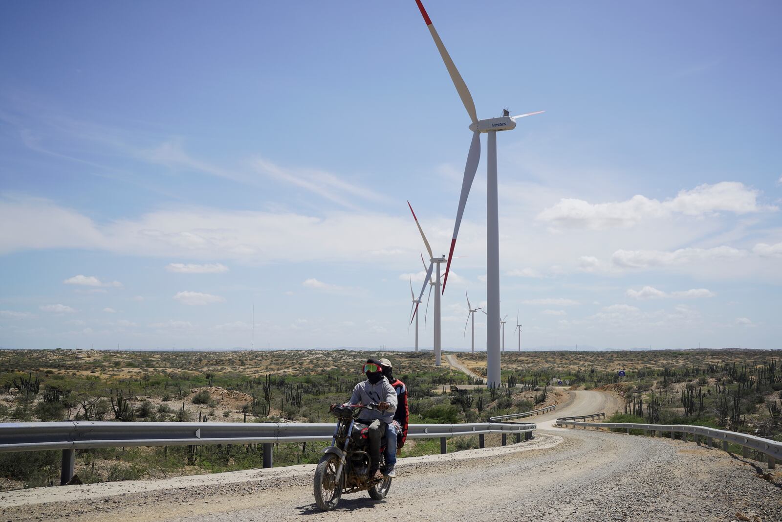 Two people on a motorcycle drive near wind turbines on the outskirts of Cabo de la Vela, Colombia, Friday, Feb. 7, 2025. (AP Photo/Ivan Valencia)