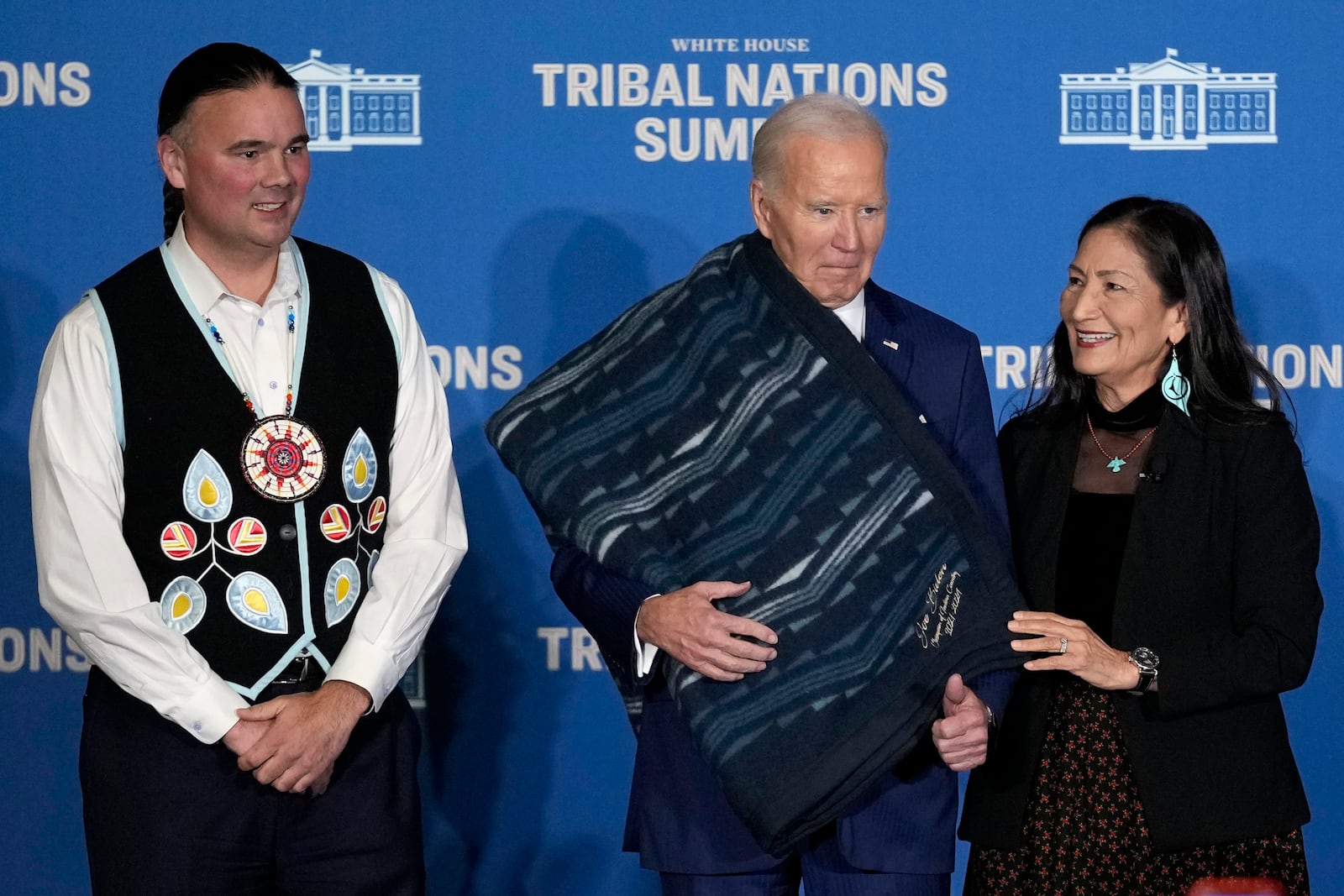 Interior Secretary Deb Haaland, right, and Bryan Newland, Assistant Secretary for Indian Affairs at the Department of the Interior, left, present President Joe Biden with an Eighth Generation blanket from a tribally-owned business, embroidered with "Joe Biden Champion for Indian Country," at the 2024 White House Tribal Nations Summit at the Department of the Interior in Washington, Monday, Dec. 9, 2024. (AP Photo/Susan Walsh)