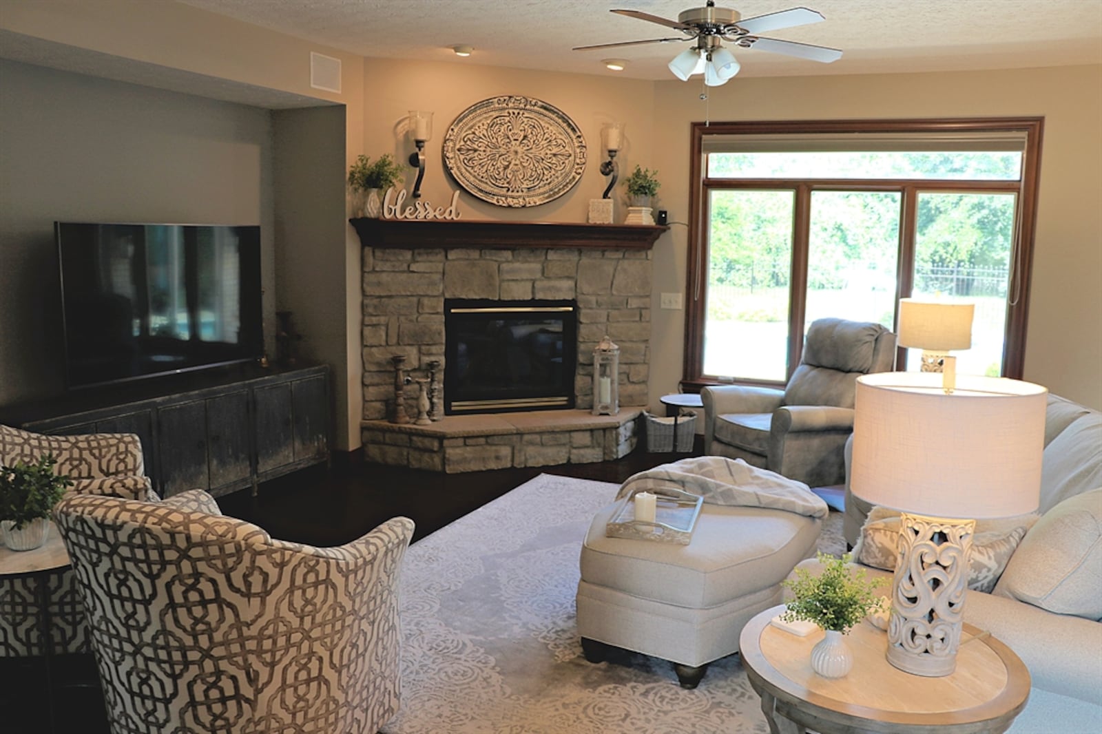 A stone gas fireplace is tucked within a corner of the great room. The fireplace has a raised stone hearth and wood-beam mantel. A furniture nook has built-in speakers above. Triple windows with a transom provide a view of the backyard pool deck.