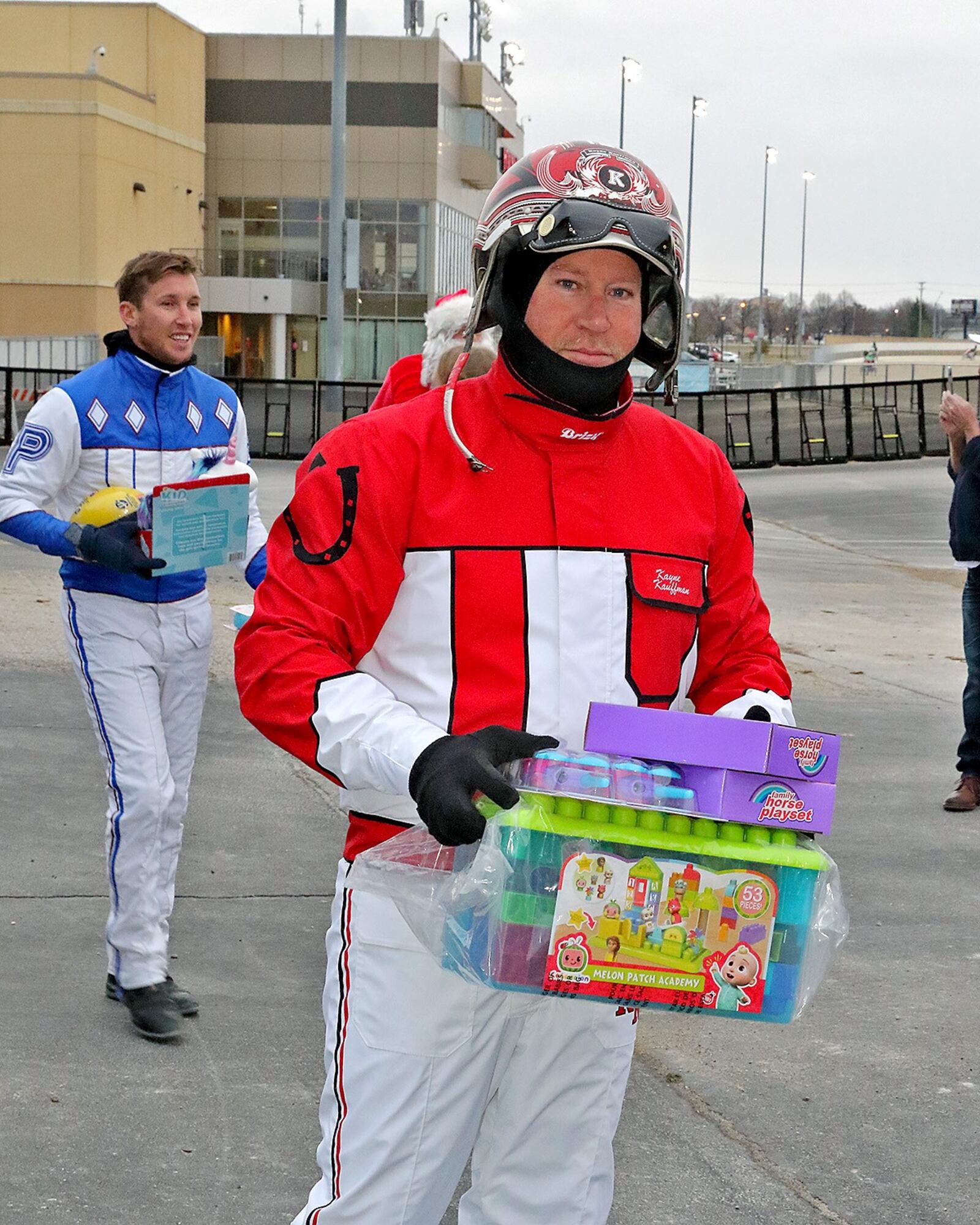 Harness drivers Kayne Kauffman (red silks) and Chris Page (blue) carry some of the many Christmas presents horsemen at Dayton Raceway donated to the annual Christmas from the Firehouse drive put on by Dayton Firefighters Union Local 136. The presents were loaded onto a waiting firetruck at the track eight days ago and were given out to local families and children in need on Christmas Eve.  (Brad Conrad/ Conrad Photos)