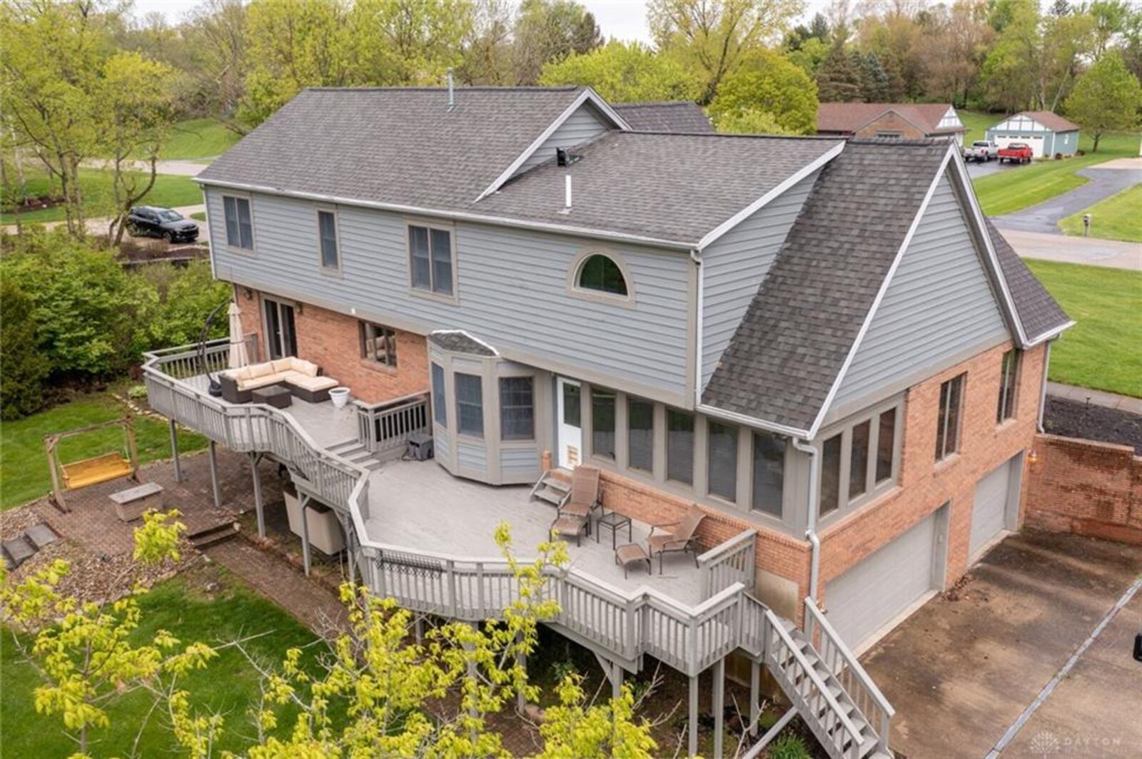 The rear of the home has a two-tiered wood deck with railings and a lower-level paver patio. The lot is wooded with a stream.