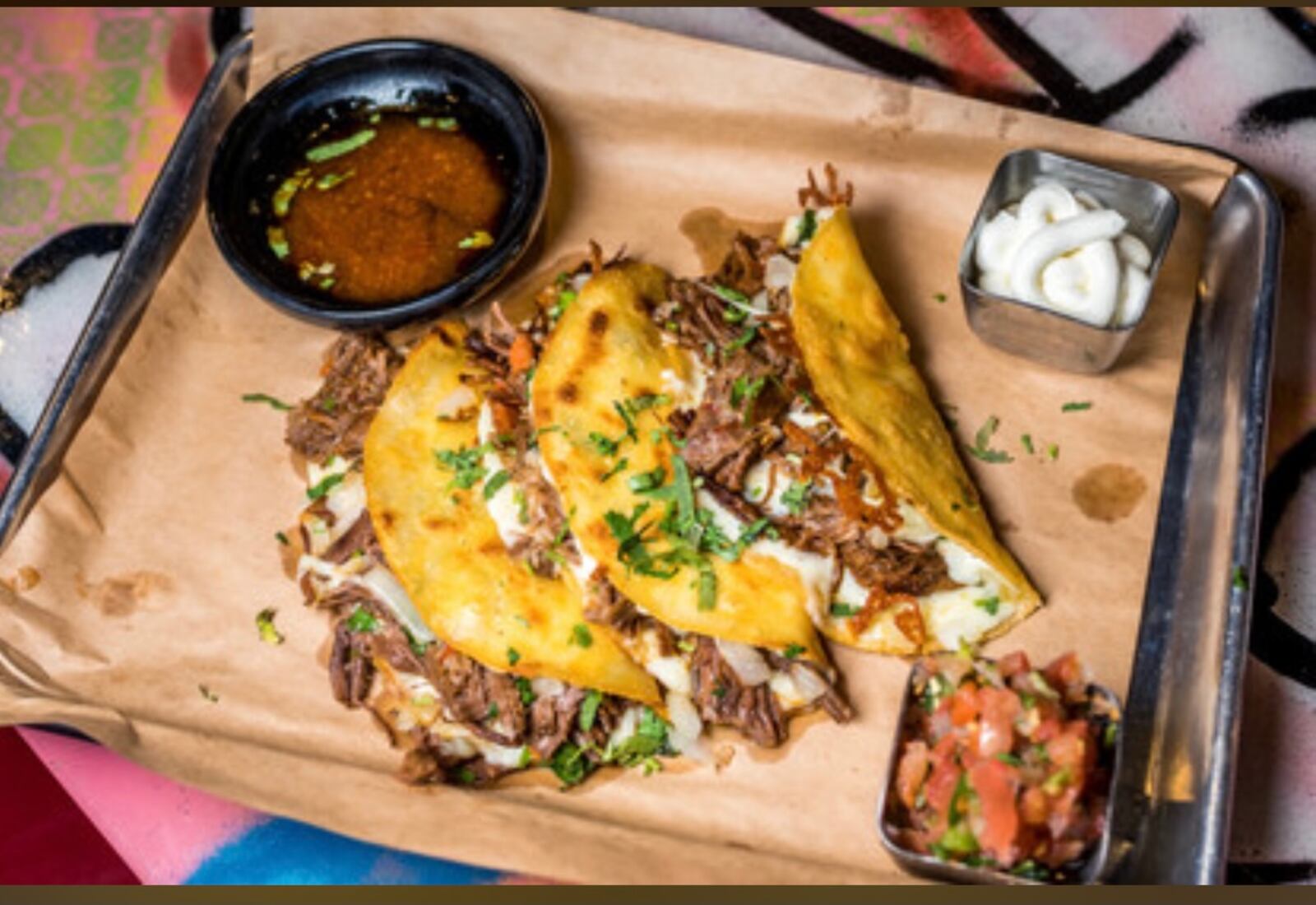 The Birria, three Slow-Braised Beef with Guajillo Chilies, Oaxaca Cheese, Cilantro, Onion, Dipping Consommé.