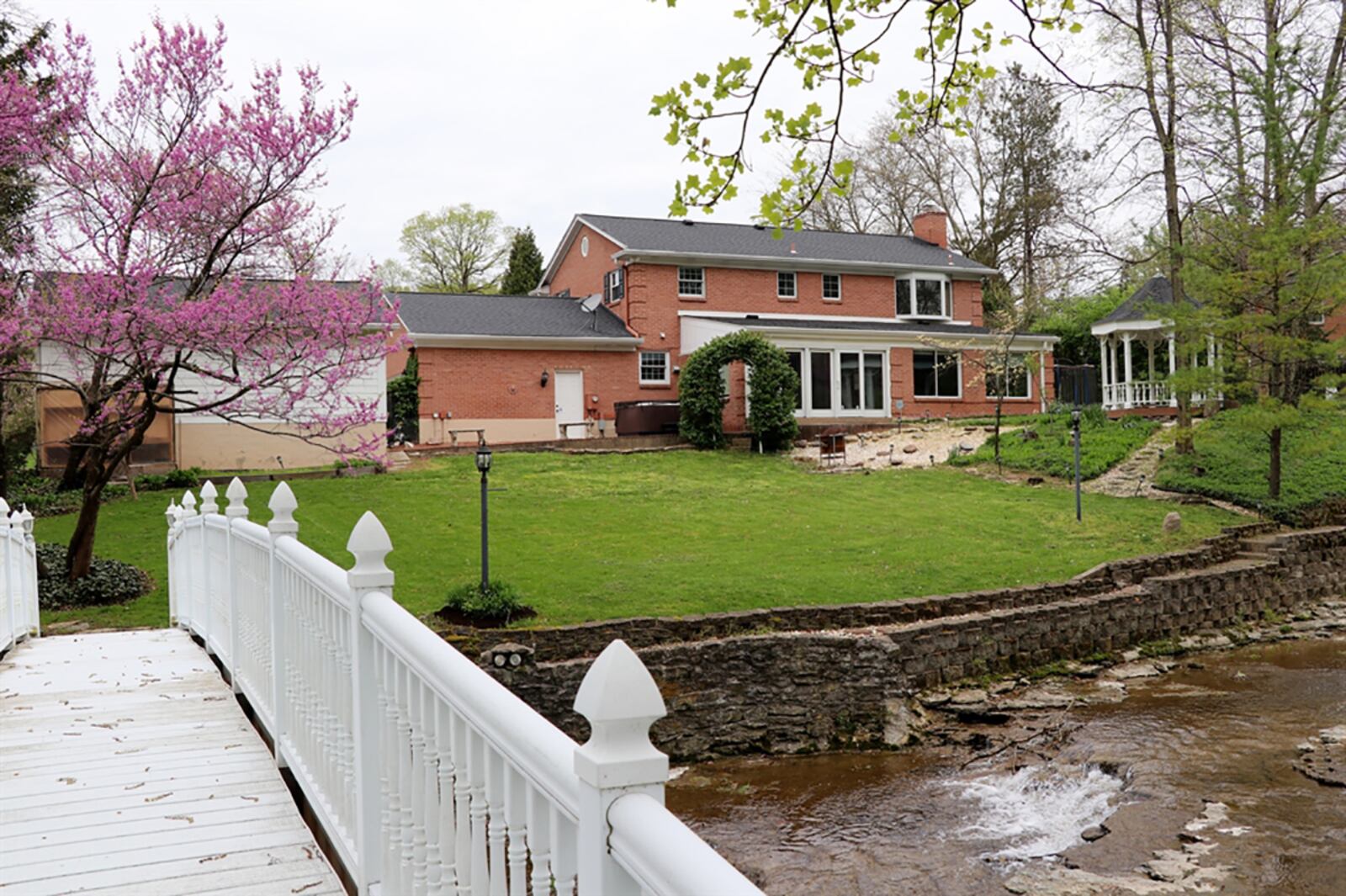 Flowering trees and evergreen trellises accent the soft rolling hills of the back yard where stone paths lead to a paver-brick patio, a wooden gazebo and a wooden park bench. A keystone-lined stream with rock waterfalls winds through the property and a white vinyl bridge arches over the stream to the wooded property. CONTRIBUTED PHOTO BY KATHY TYLER