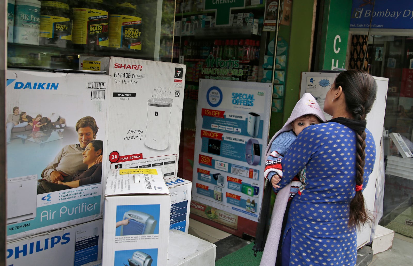 FILE- A woman carries an infant outside a shop selling air purifiers at a market place in New Delhi, India, on Nov. 14, 2017. (AP Photo/Altaf Qadri, File)