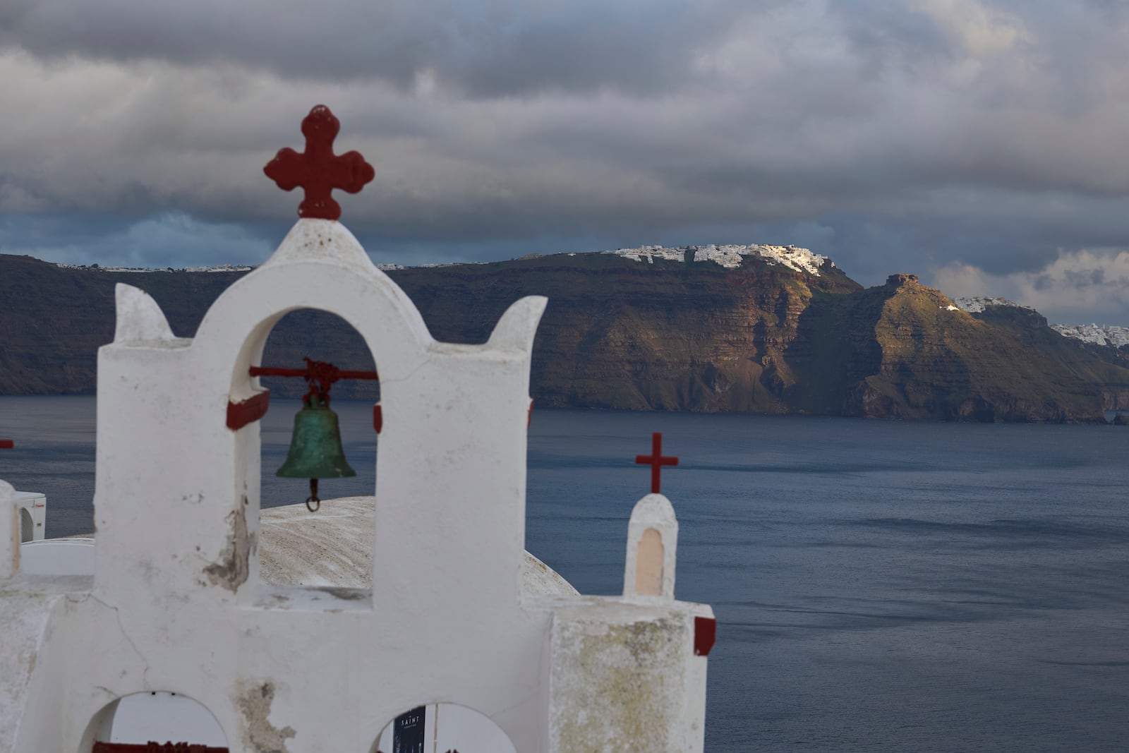 A bell towel of an Orthodox church in the town of Oia on the earthquake-struck island of Santorini, Greece, as the main town of Fira is litted by the sun in the background on Tuesday, Feb. 4, 2025. (AP Photo/Petros Giannakouris)