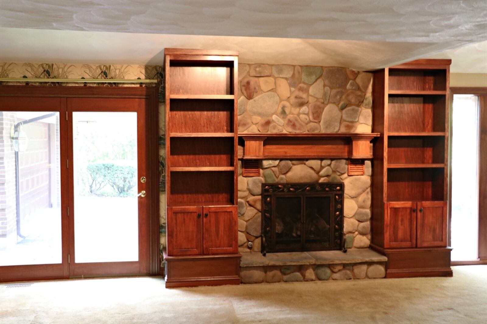 A stone gas fireplace, centered between the windows and the patio doors, has a raised hearth and wood-beam mantel. Bookcases with cabinetry flank the fireplace. 