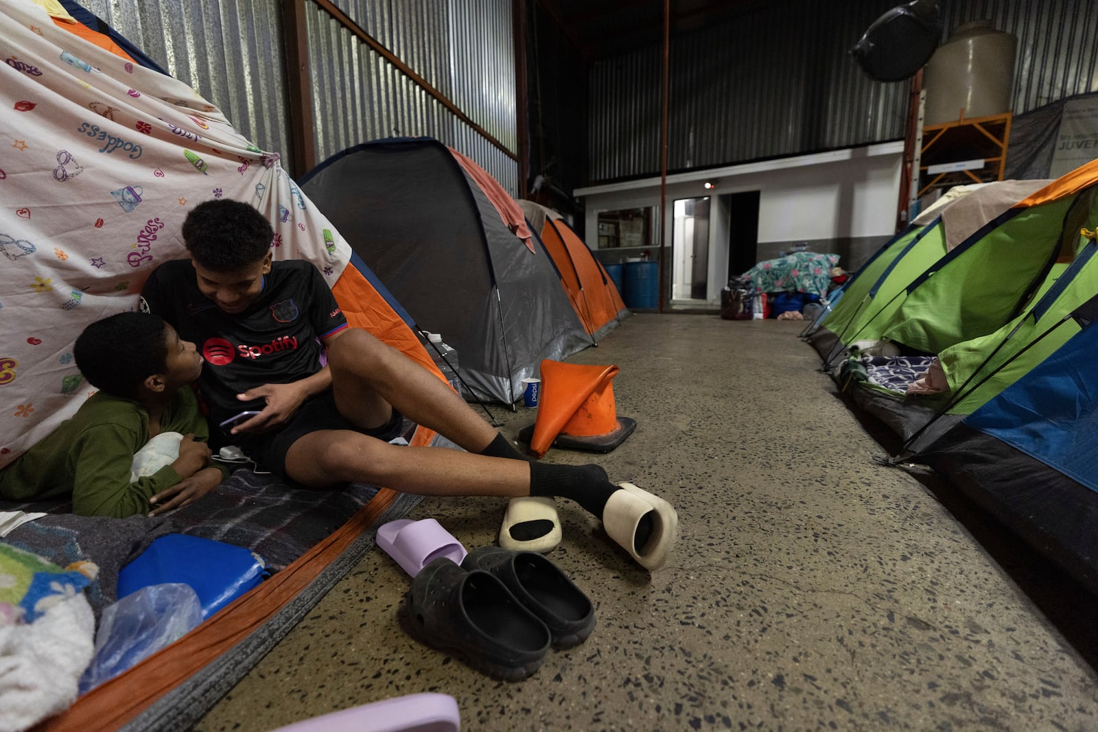 Mickel Rodriguez, 12, of Venezuela, left, watches videos on a phone with a family friend who is traveling with his family as they rest in their tent at a migrant shelter in Tijuana, Mexico, Sunday, Feb. 2, 2025. (AP Photo/Gregory Bull)