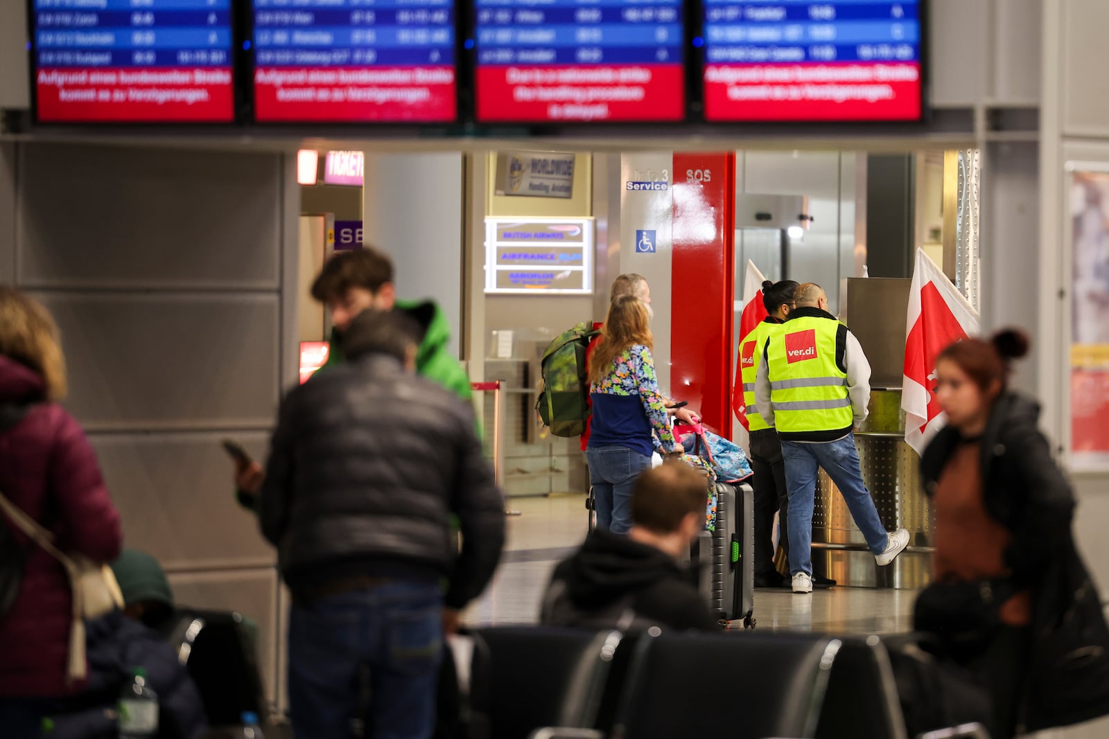 Airport employees wearing high-visibility vests from the Verdi union stand in the departures area at Dusseldorf Airport, Germany while travelers wait for their flight under the display boards Monday, March 10, 2025. (Christoph Reichwein/dpa via AP)