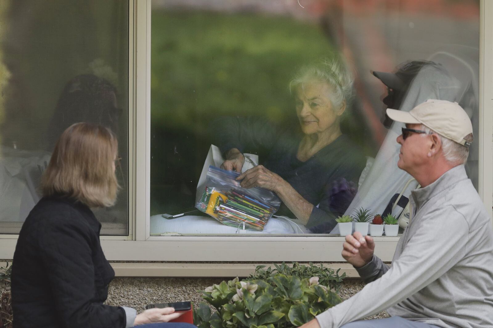 Judie Shape, center, who has tested positive for the new coronavirus, but isn’t showing symptoms, opens a care package of art supplies from her daughter Lori Spencer, left, and her son-in-law Michael Spencer, Tuesday, March 17, 2020, as they talk on the phone and look at each other through a window at the Life Care Center in Kirkland, Wash., near Seattle. In-person visits are not allowed at the nursing home, which is at the center of the outbreak of the new coronavirus in the United States. (AP Photo/Ted S. Warren)