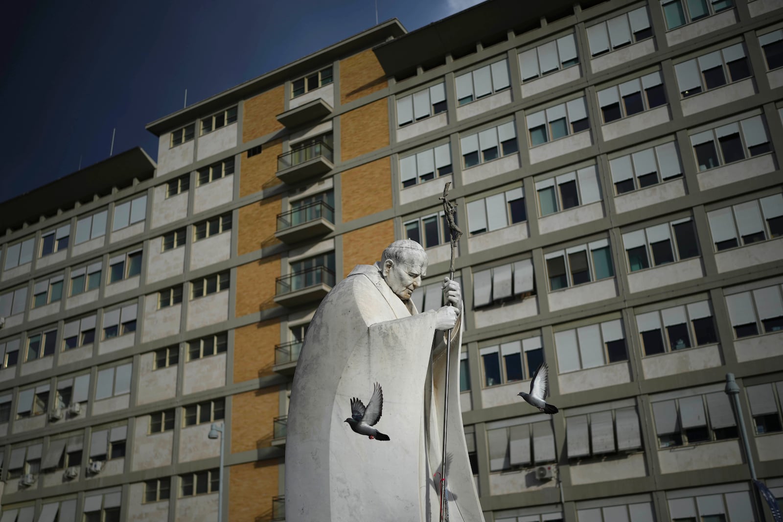 A marble statue of late Pope John Paul II is backdropped by the Agostino Gemelli Polyclinic in Rome, Saturday, Feb. 15, 2025, where Pope Francis was hospitalised Friday after a weeklong bout of bronchitis worsened and is receiving drug therapy for a respiratory tract infection. (AP Photo/Alessandra Tarantino)