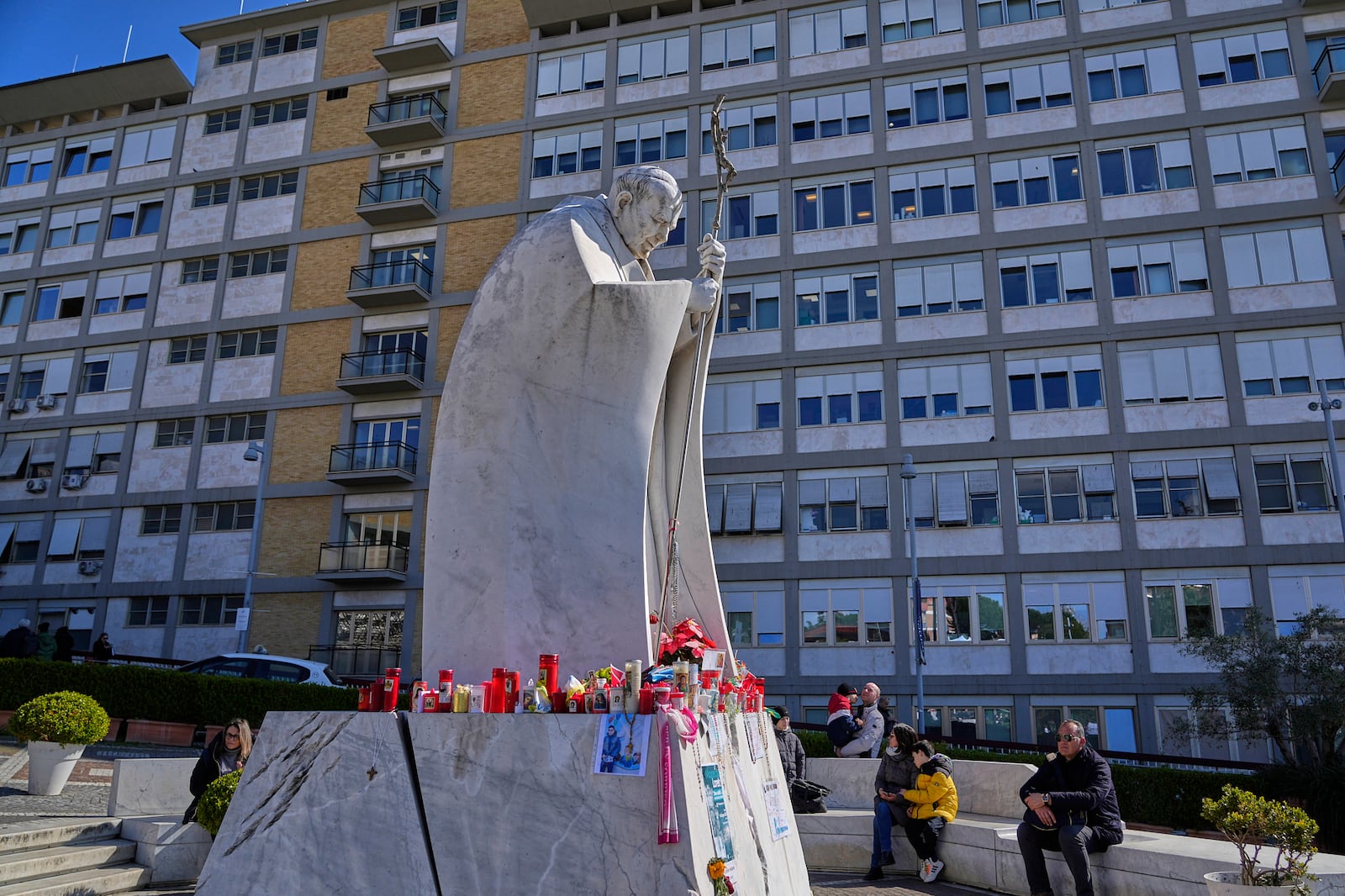 People sit by the statue of Pope John Paul II set outside the Agostino Gemelli Polyclinic where Pope Francis in Rome, Tuesday, March 4, 2025. (AP Photo/Gregorio Borgia)