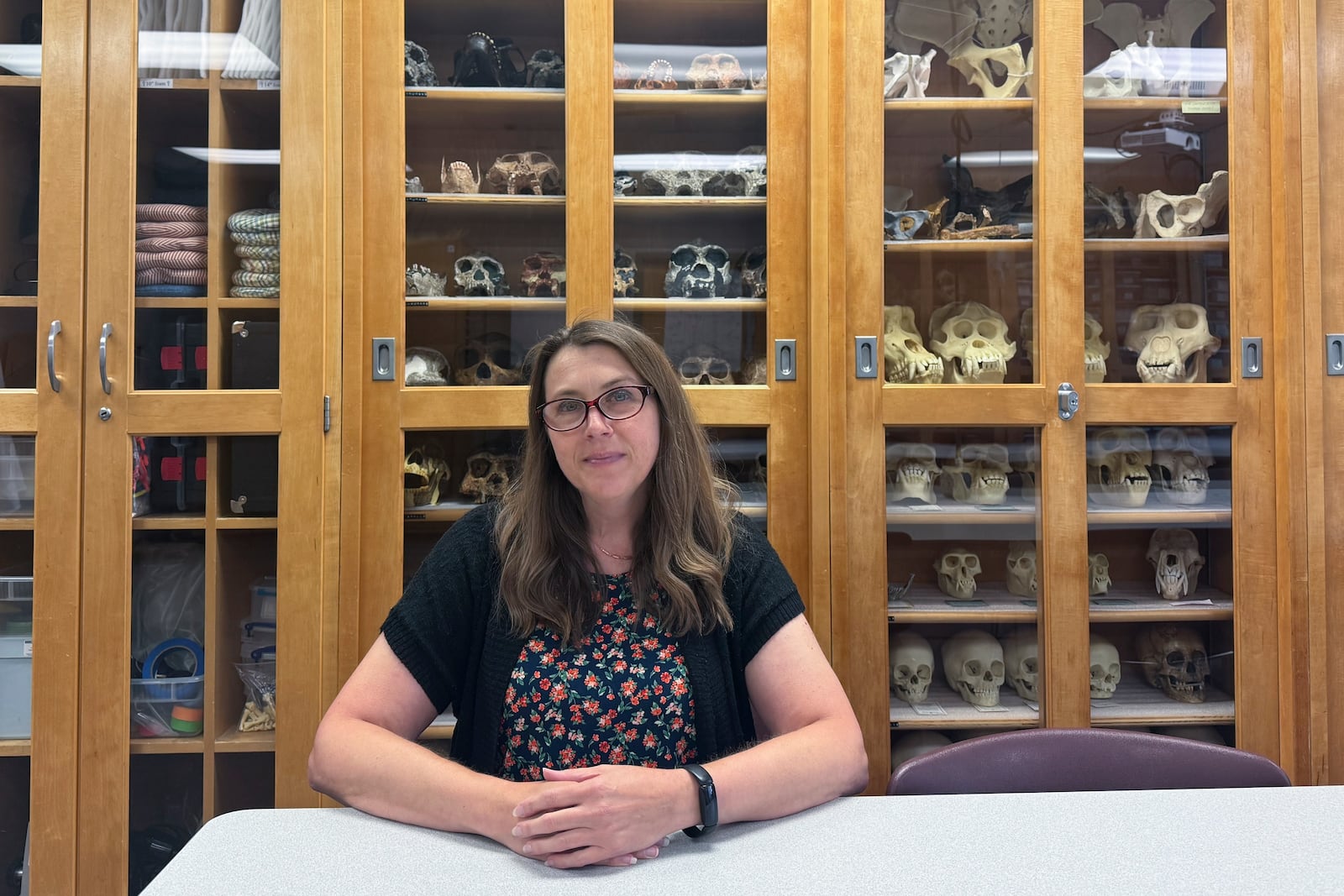 Dr. Krista Latham, the director of the Human Identification Center at the University of Indianapolis, sits at a table in front of the center's cabinets containing replicas of fossil finds of ancient humans , Aug. 1, 2024, in Indianapolis. (AP Photo/Rick Callahan)