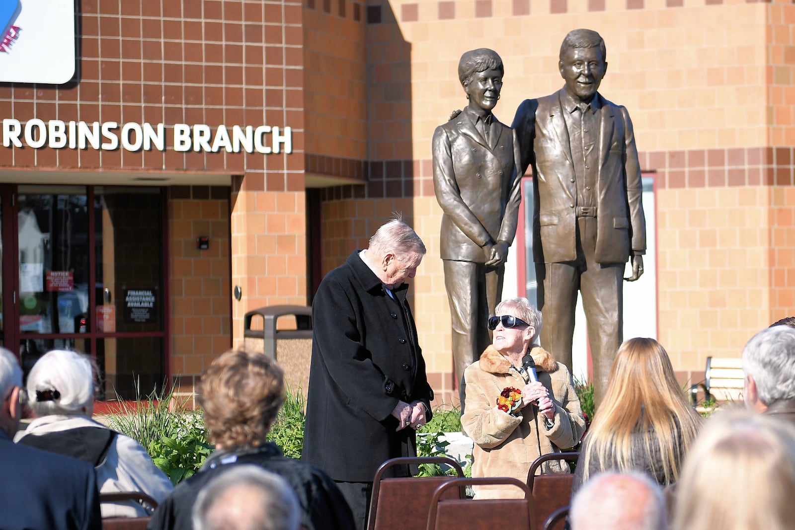 Thom Robinson (standing) and Pat Robinson (seated with microphone) address those attending the unveiling of sculptures of the Troy philanthropists at the Miami County YMCA Robinson Branch on Nov. 5. CONTRIBUTED