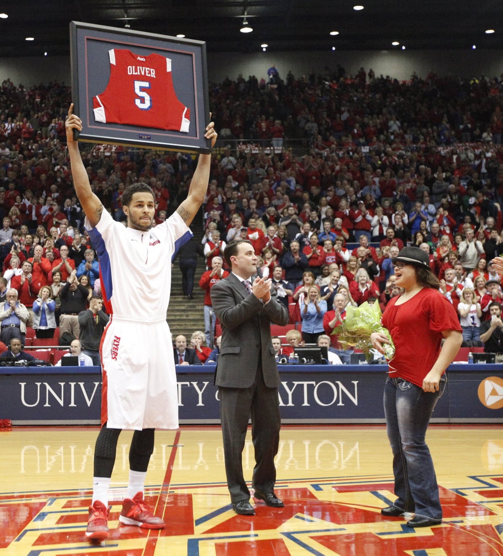 Dayton senior Devin Oliver, left, holds up a framed jersey on Senior Night as coach Archie Miller and Oliver’s sister Miya applaud before a game against Richmond on Saturday, March 8, 2014, at UD Arena. David Jablonski/Staff