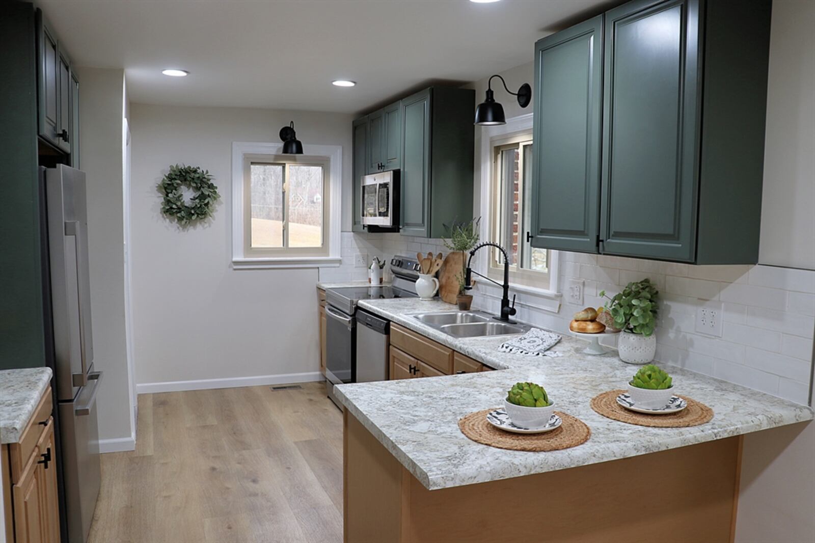 Green shaker hanging cabinets complement the lower light maple cabinets within the kitchen. A pantry cabinet surrounds the refrigerator nook, and open shelves are above the coffee station with subway tile backsplash.