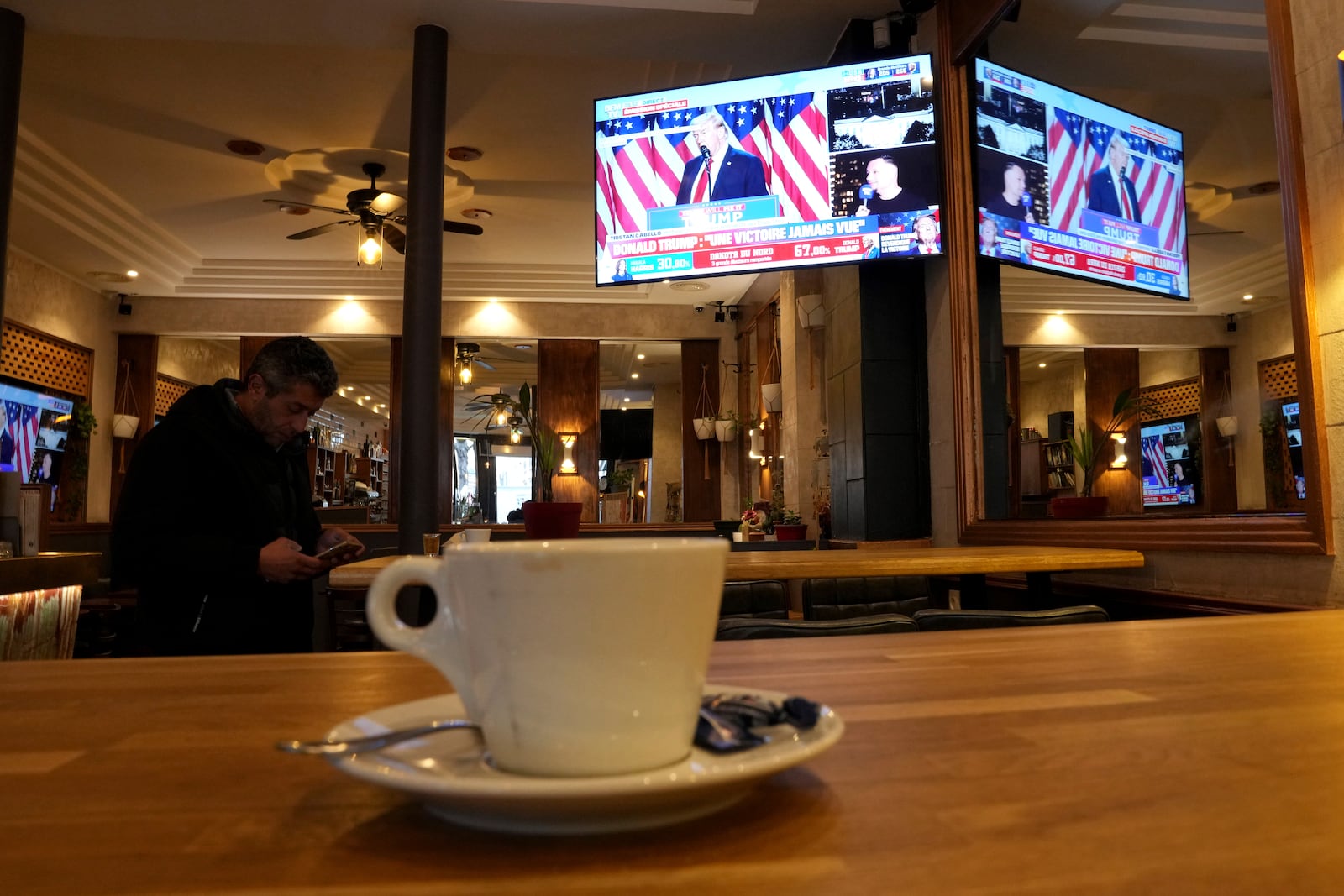 A man checks his smartphone in a cafe as a television screen shows Donald Trump,Wednesday, Nov. 6, 2024 in Paris. (AP Photo/Aurelien Morissard)