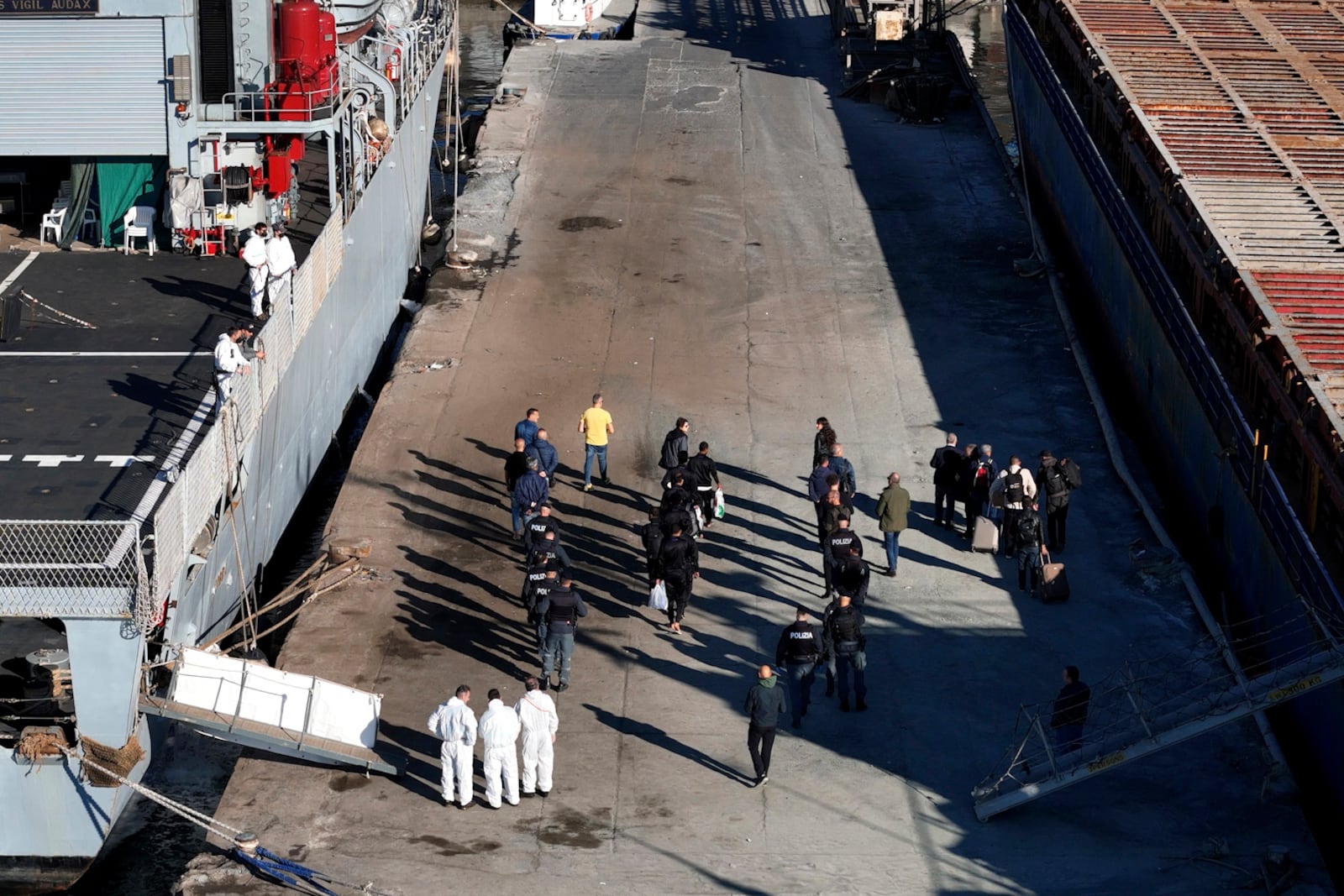 Migrants walk at the port of Shengjin, next to security forces, after they disembark from an Italian Navy ship northwestern Albania, Friday, Nov. 8, 2024, while a second group of eight migrants intercepted in international waters is processed in a reception facility despite the failure with the first group in October. (AP Photo/Vlasov Sulaj)