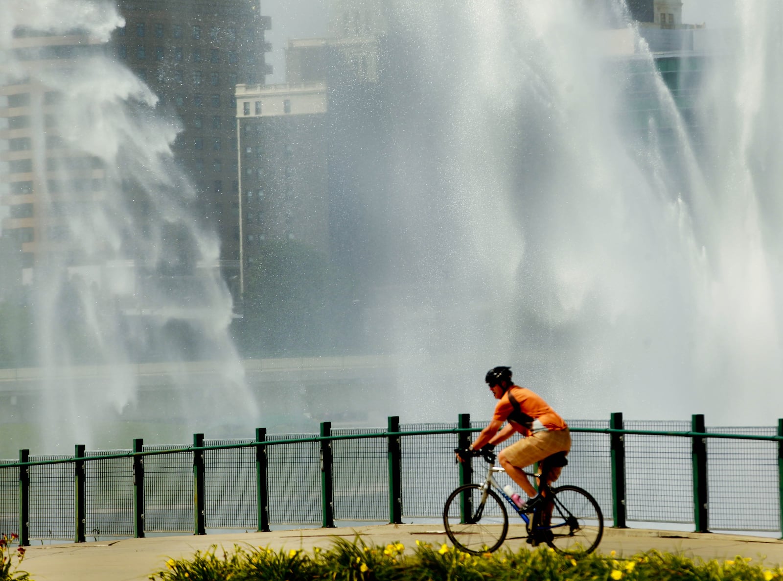 A cyclist rides along a backdrop of a noon-hour display of the spraying fountains at the Deeds Point area of Riverscape recently in downtown Dayton. JIM WITMER / STAFF