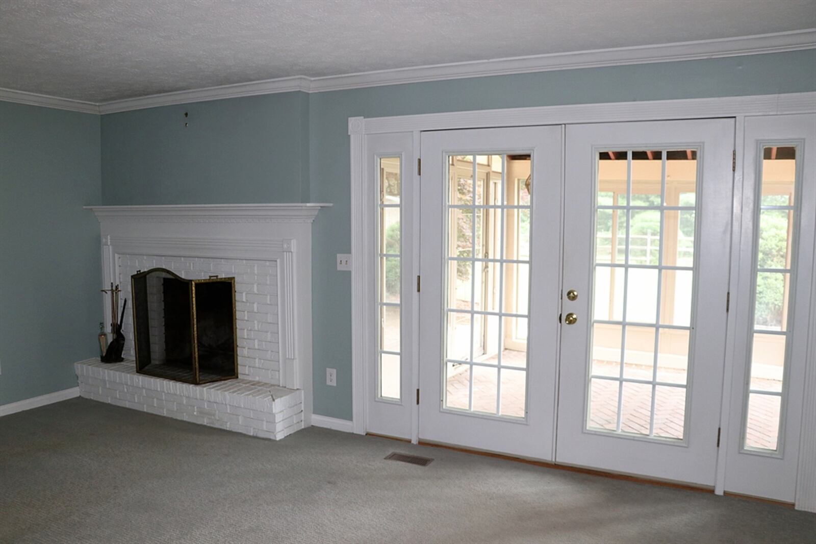 A spacious family room has a wood-burning fireplace tucked near a corner. The fireplace has a raised brick hearth and dentil wood mantel. Next to the fireplace, French doors allow access to an enclosed rear porch.