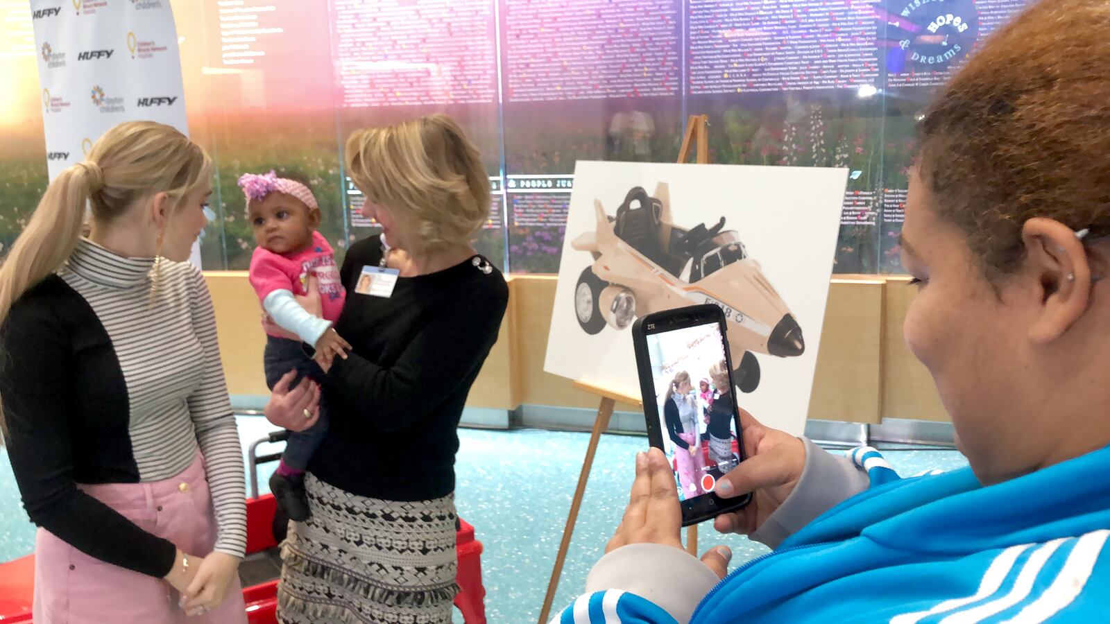 Meg Donnelly, star of  ABC’s  “American Housewife” and the popular Disney Channel movie “Zombies” smiles at 5-month-old Rai’Mia Bass being held by Julie Cannon of Dayton Children’s Hospital.  Rai’Mia had just finished a chemotherapy treatment. The baby’s mother, Brittany Bass, records a message for her 6-year-old daughter, a Donnelly fan. Donnelly visited Dayton Children’s as part of the announcement of Huffy’s donation to  Dayton Children’s and other hospitals in the Children’s Miracle Network. Photo by Amelia Robinson