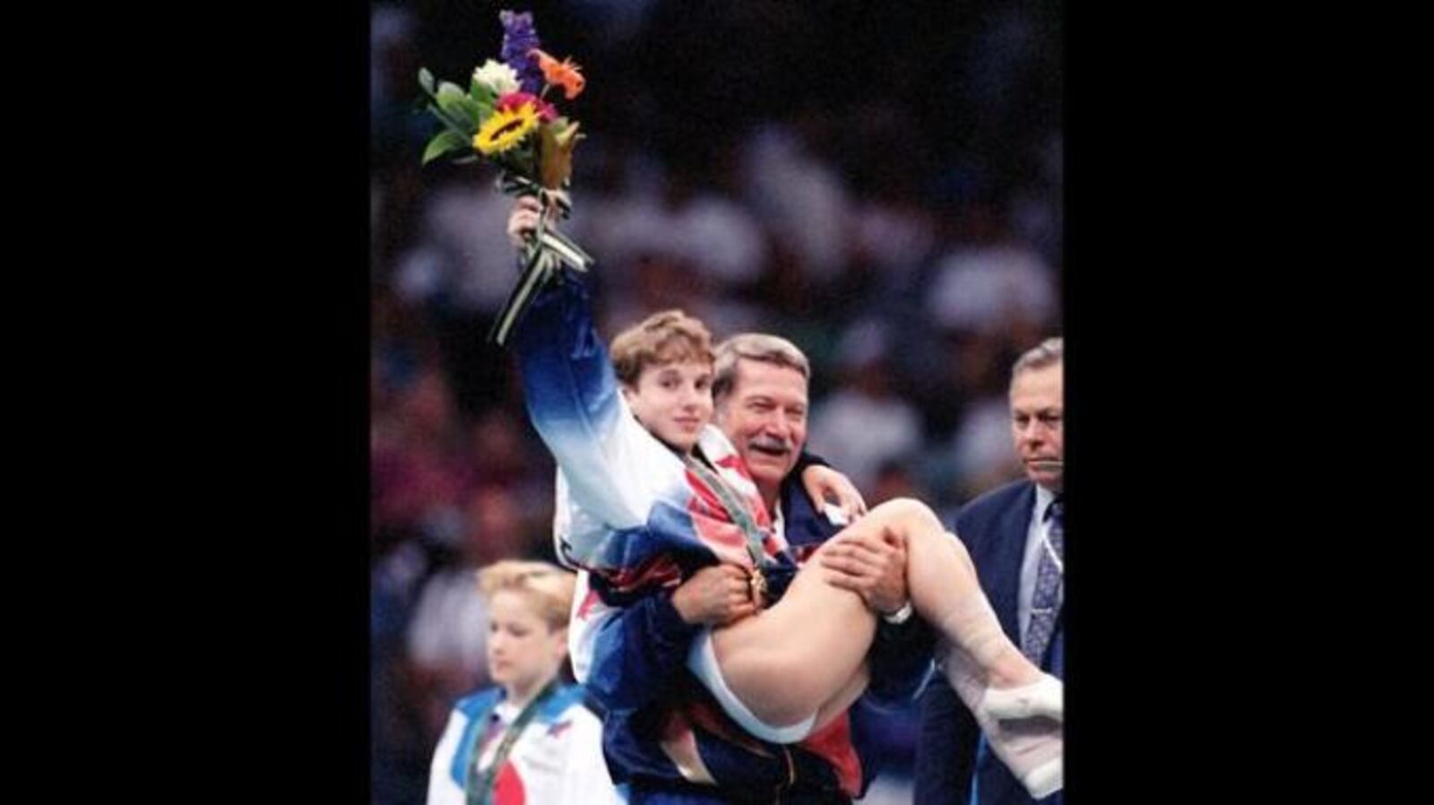 Kerri Strug hoists her flowers to the cheering crowd as she is carried from the podium by team coach, Bela Karolyi at the Georgia Dome on Tuesday, July 23, 1996. (Photo by Skip Peterson / The Atlanta Journal-Constitution)